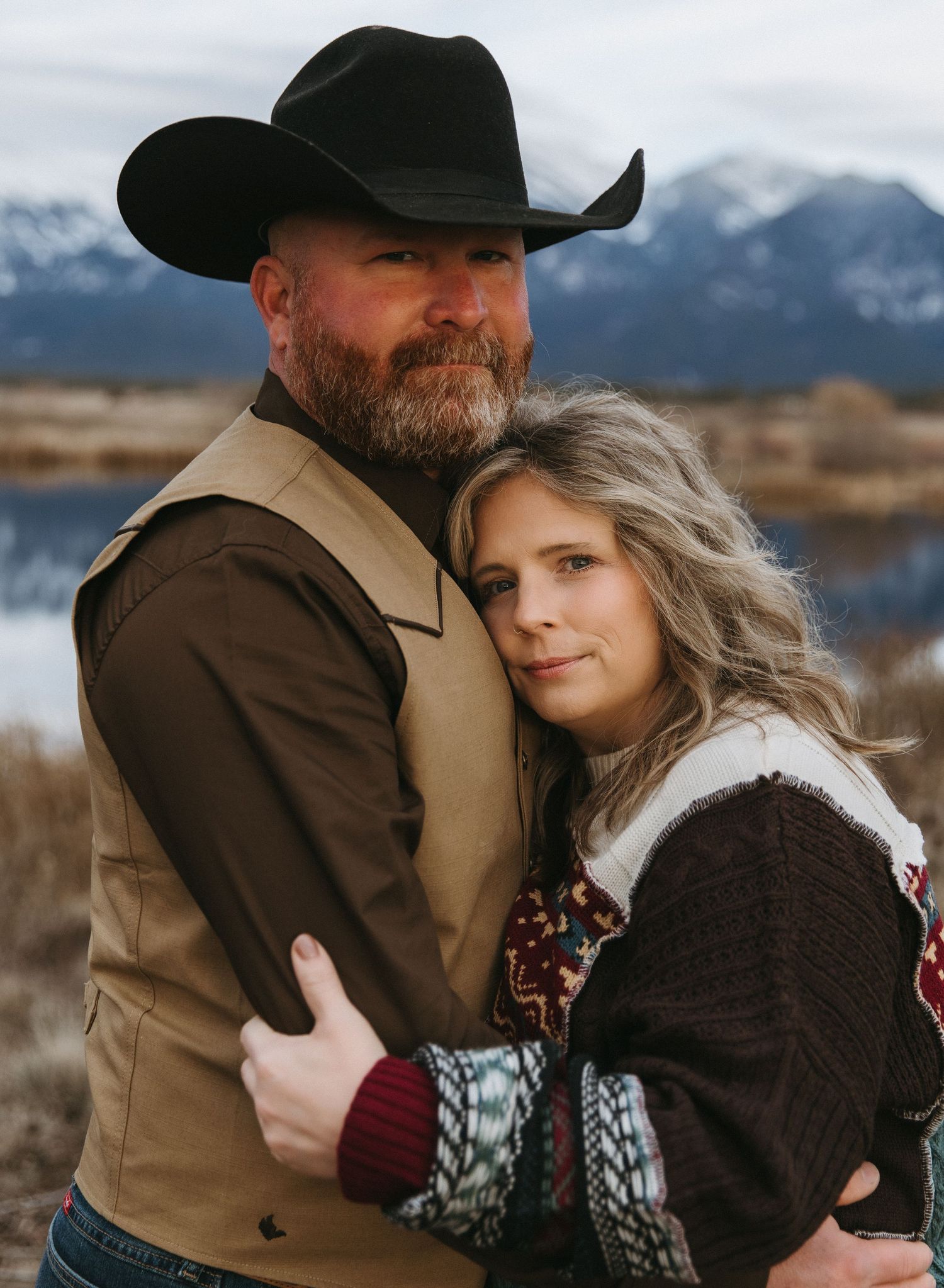 A couple embraces in front of snowy mountains wearing winter clothing and western attire.