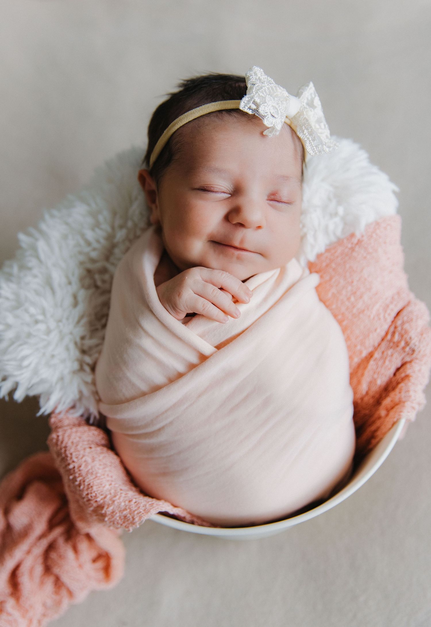 Swaddled baby sleeping in bowl with white bow headband and coral accents.