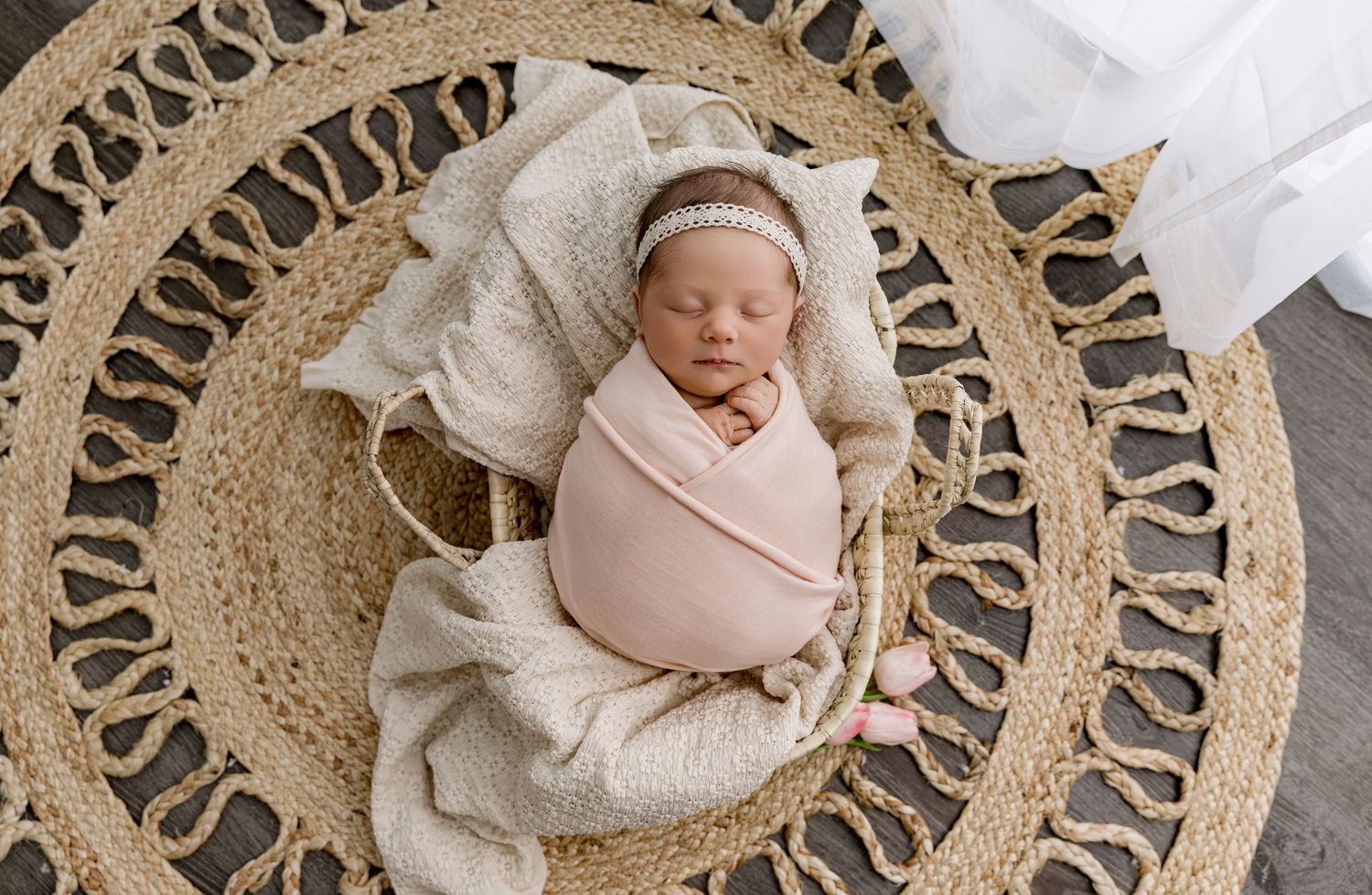 Newborn baby swaddled in pink wrap sleeping peacefully in a woven basket on a round jute rug.