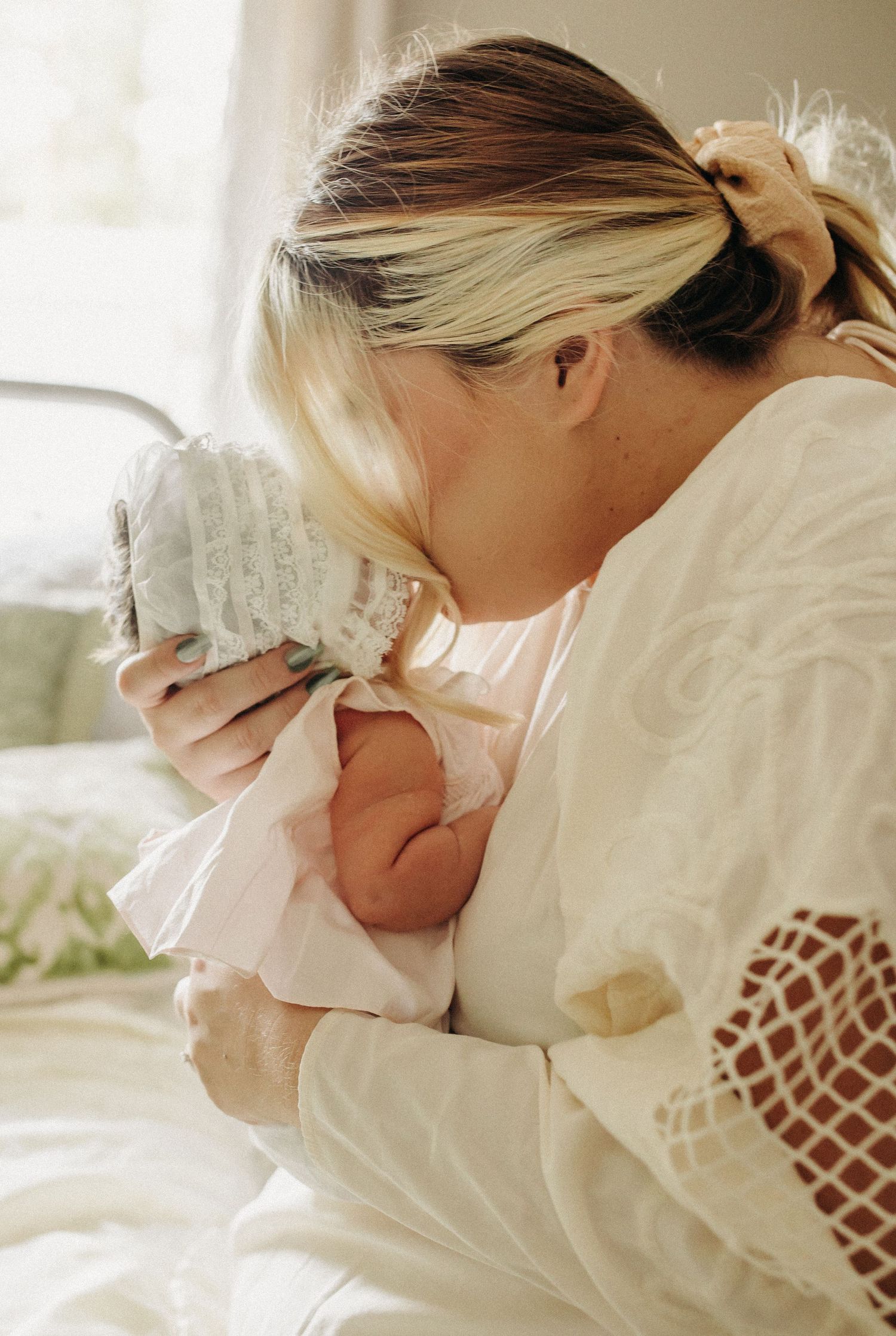 A mother gently kisses her babies forehead in the beautiful dim lit bedroom during their in home session