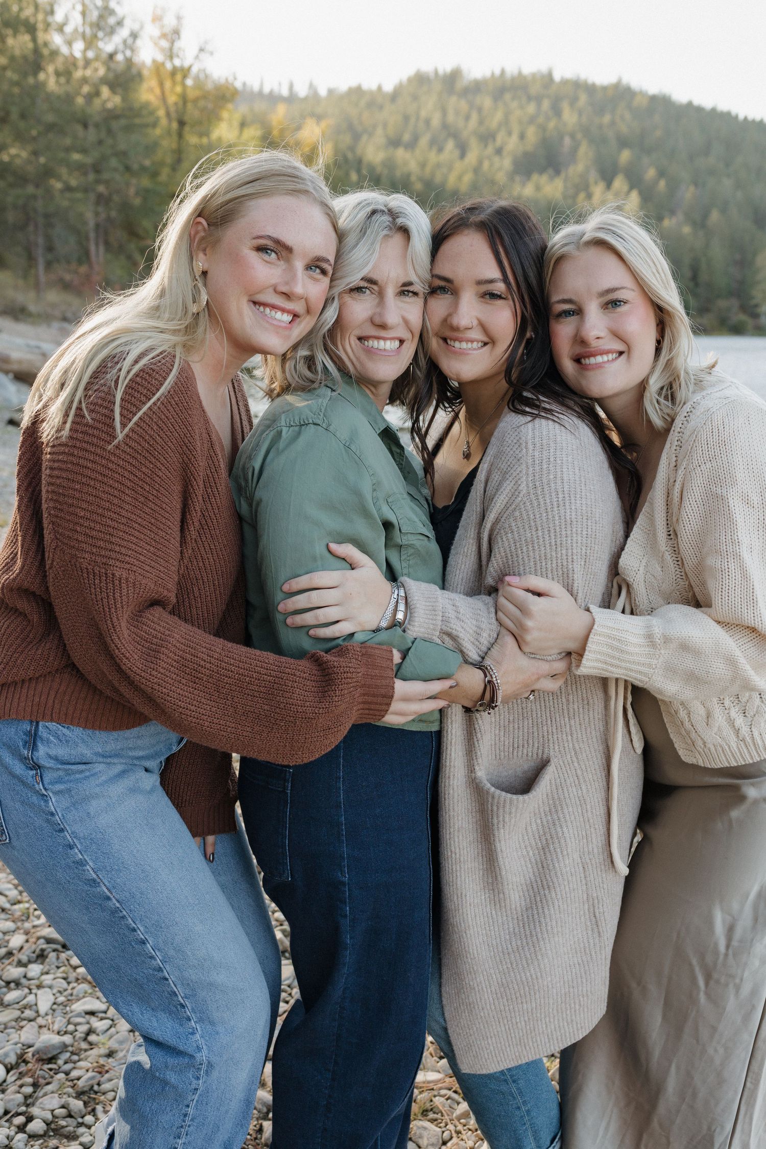 Four women in cozy sweaters embrace and smile on a rocky beach with mountains behind them.