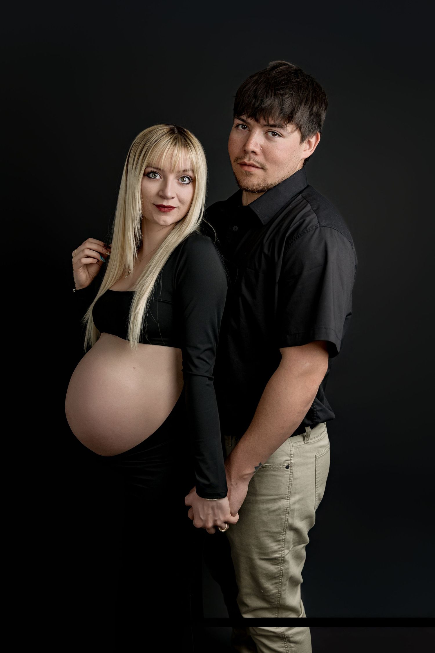 A couple poses against a dark background in a series of intimate studio portraits with moody lighting.
