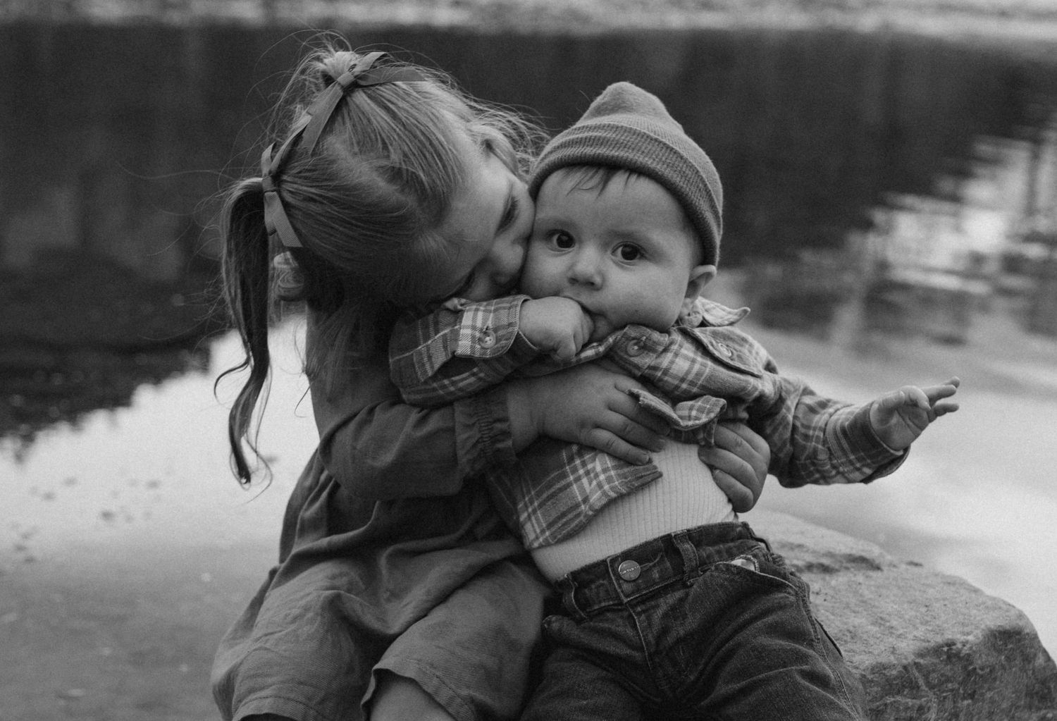 A tender black and white moment captures two small children, one giving the other an affectionate kiss by a pond.