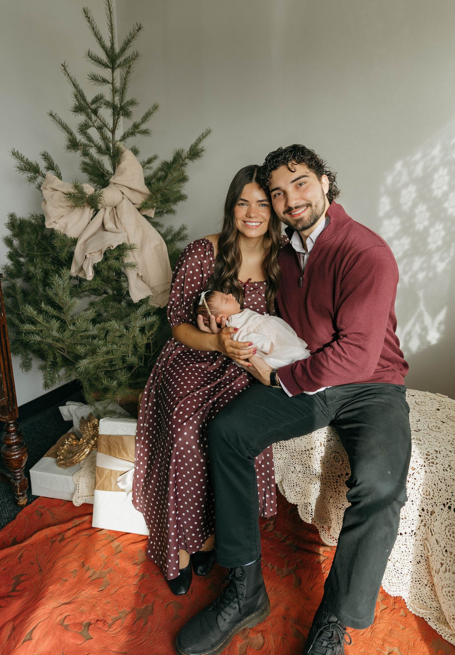 A couple poses together with their baby in front of a Christmas tree for a cozy holiday photoshoot.