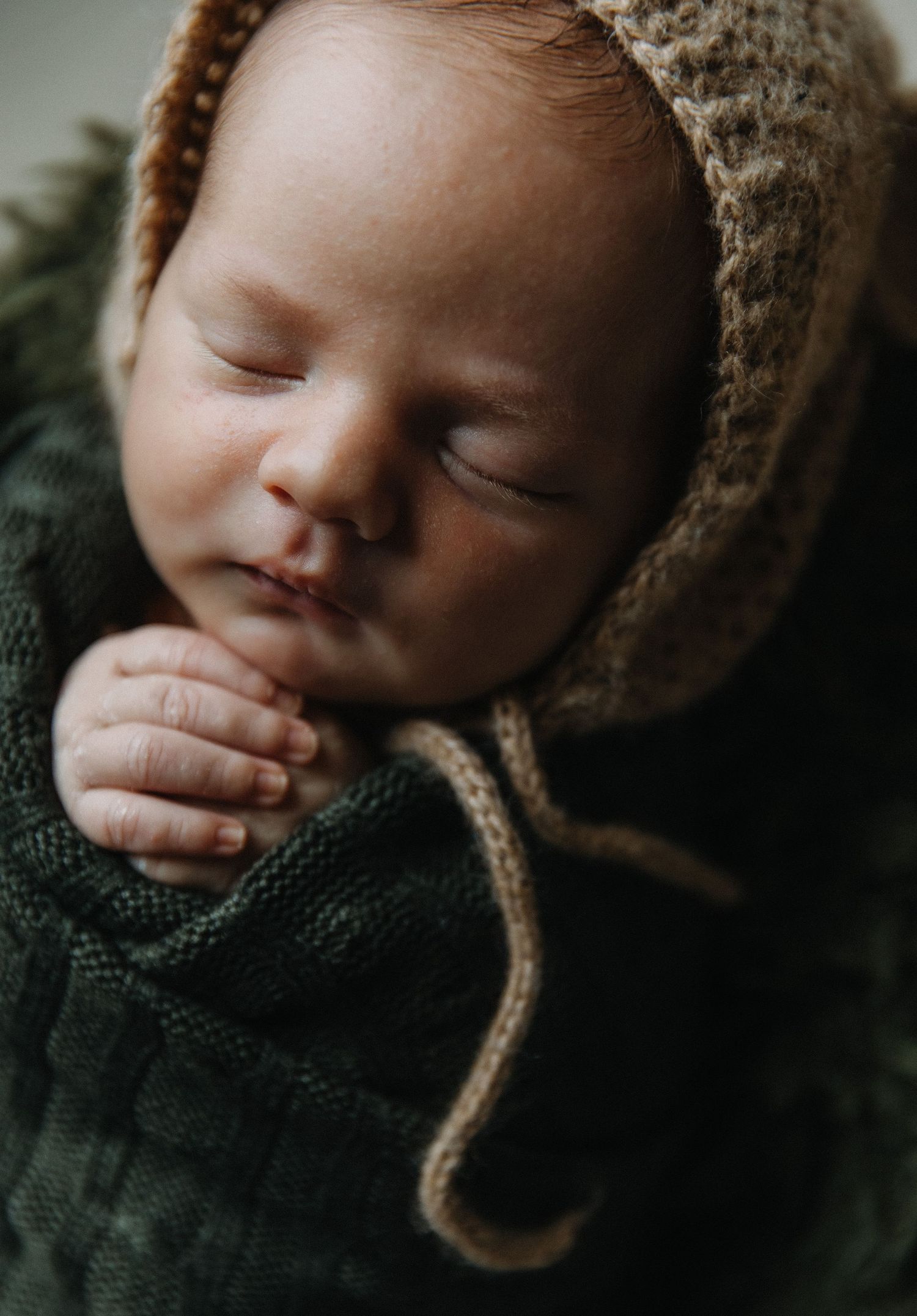 Peaceful sleeping baby wearing brown bonnet and green knit sweater in studio setting.