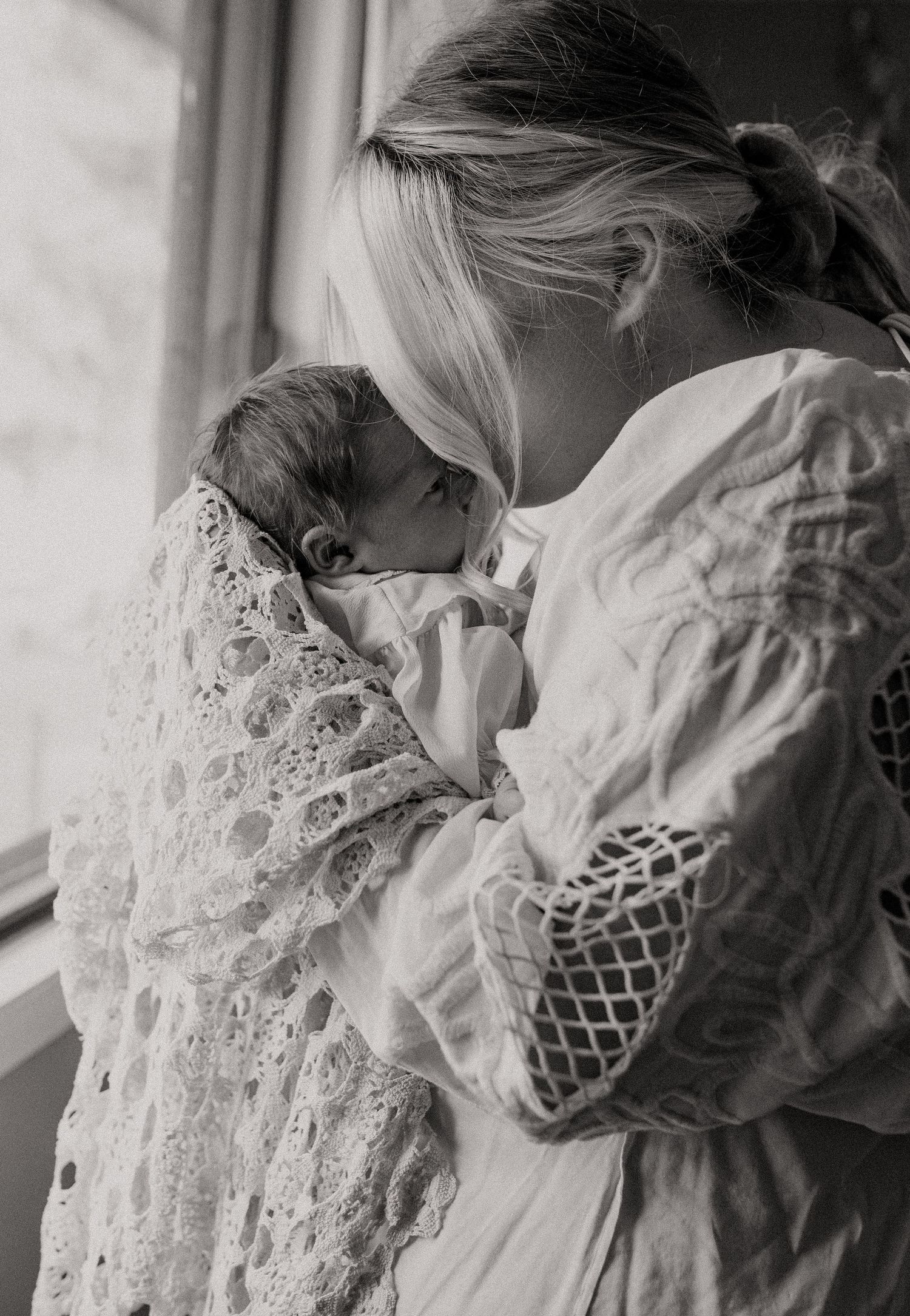 A mother gently kisses her babies forehead in the beautiful dim lit bedroom during their in home session