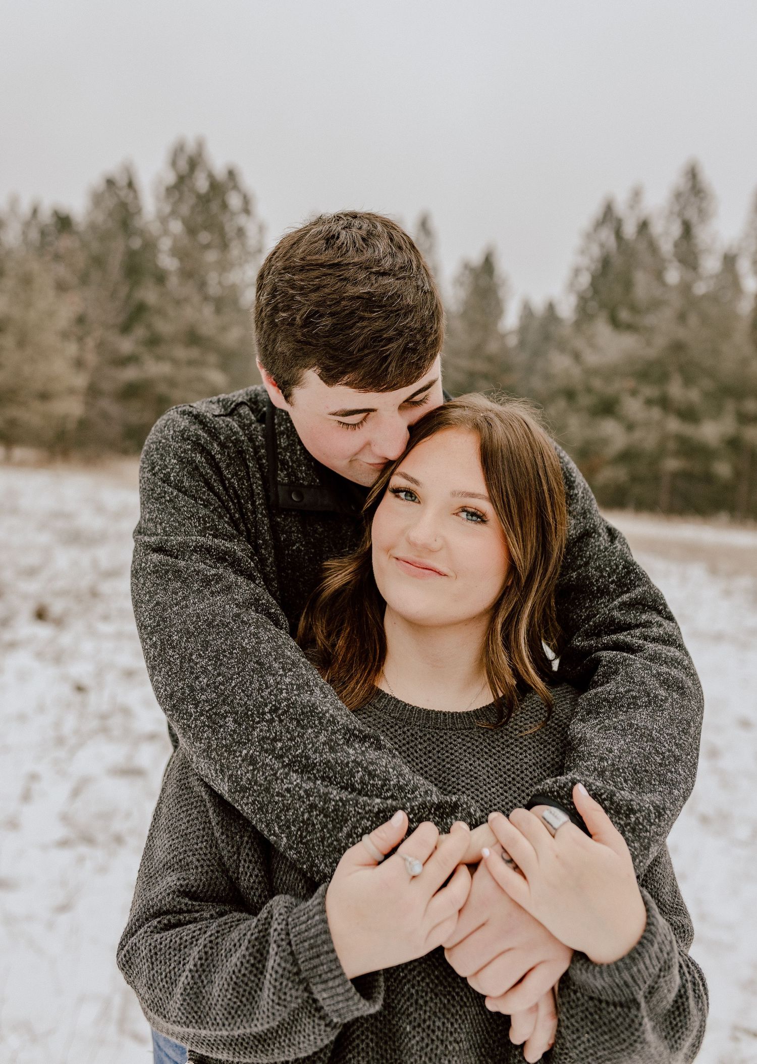 A cozy winter engagement photo of a couple embracing in the snow with pine trees in background.