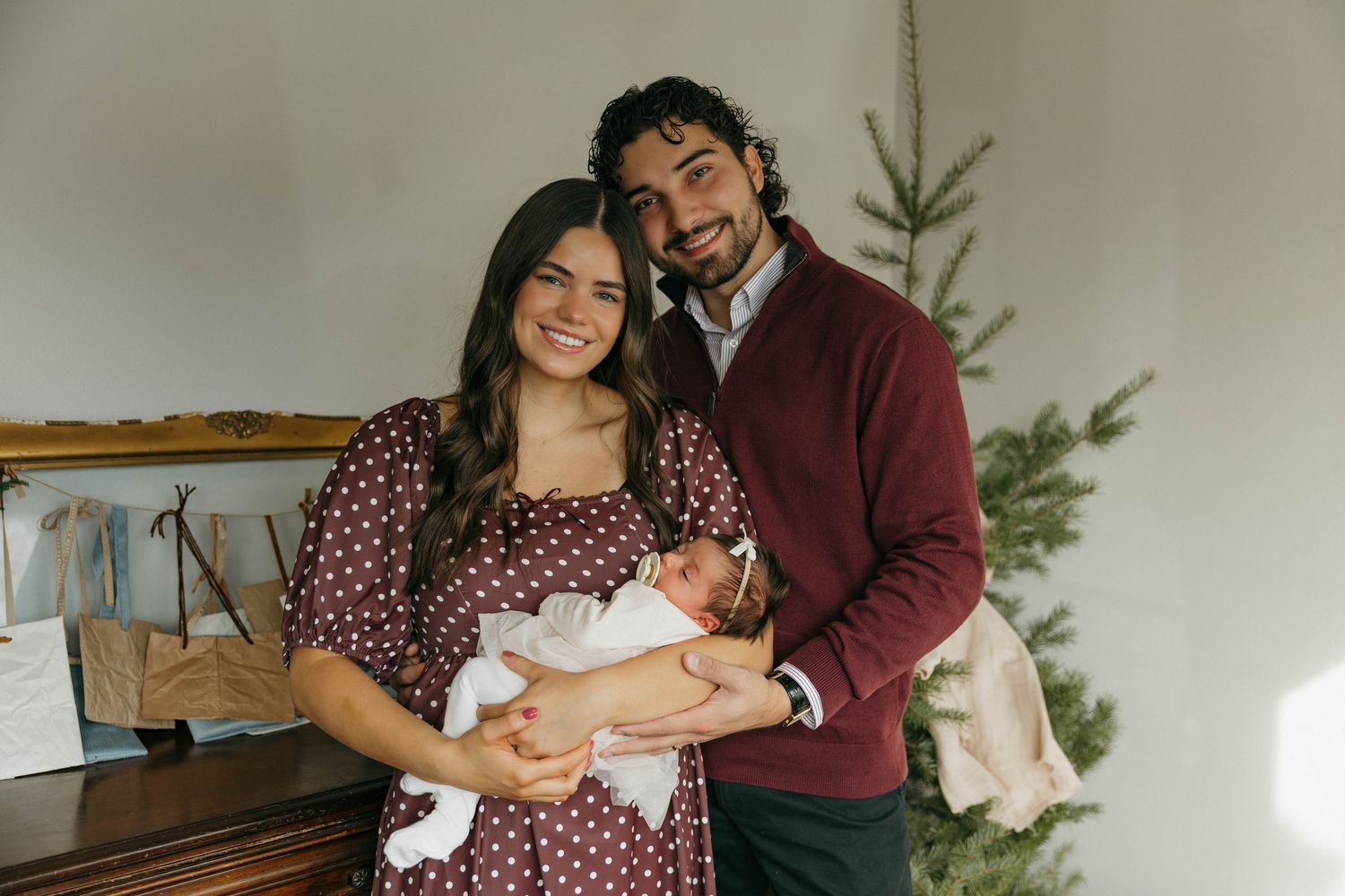 A couple in burgundy and floral print outfits pose together in a cozy indoor setting with holiday decorations.