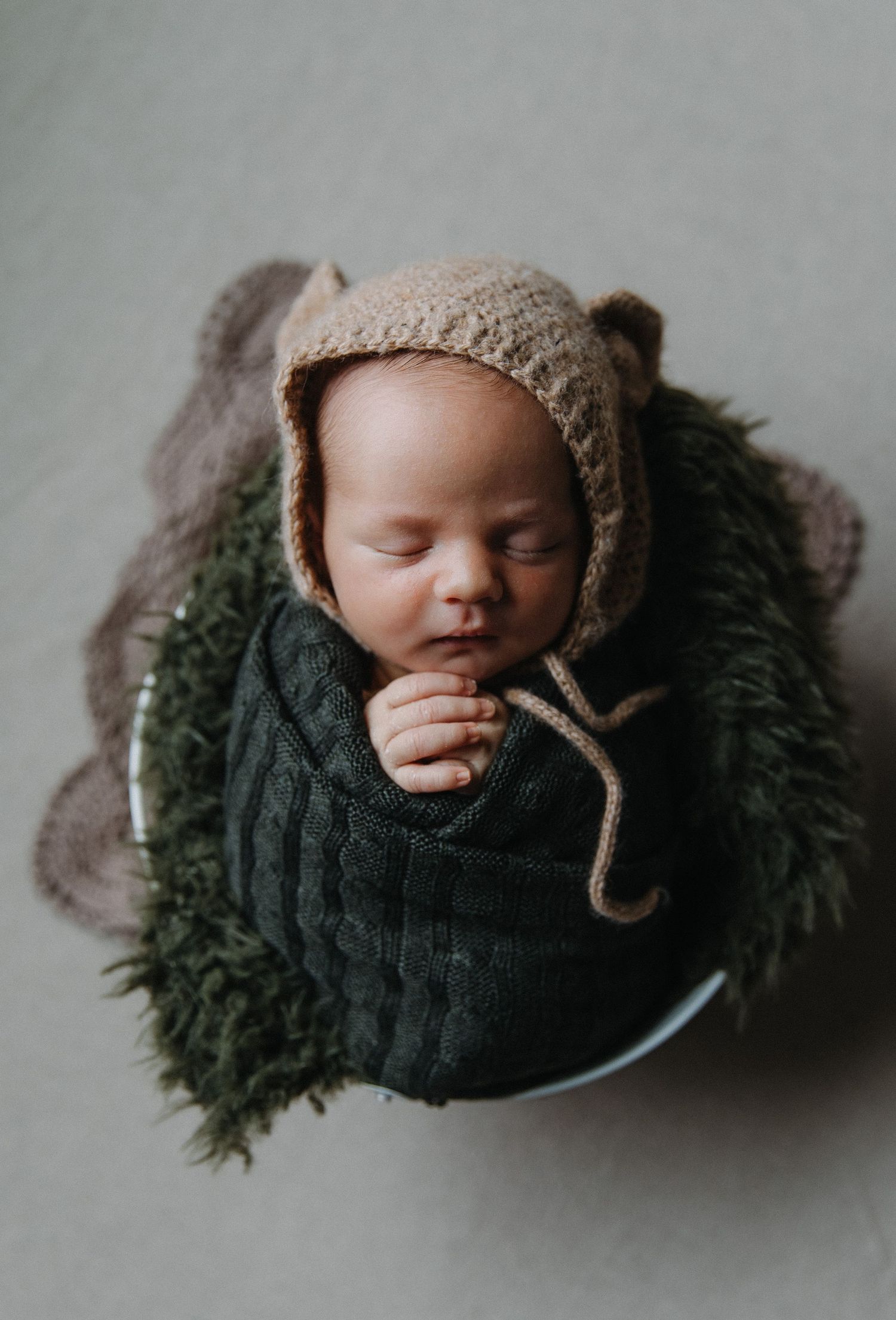 Sleeping newborn in knit bear bonnet and sweater on green textured backdrop.