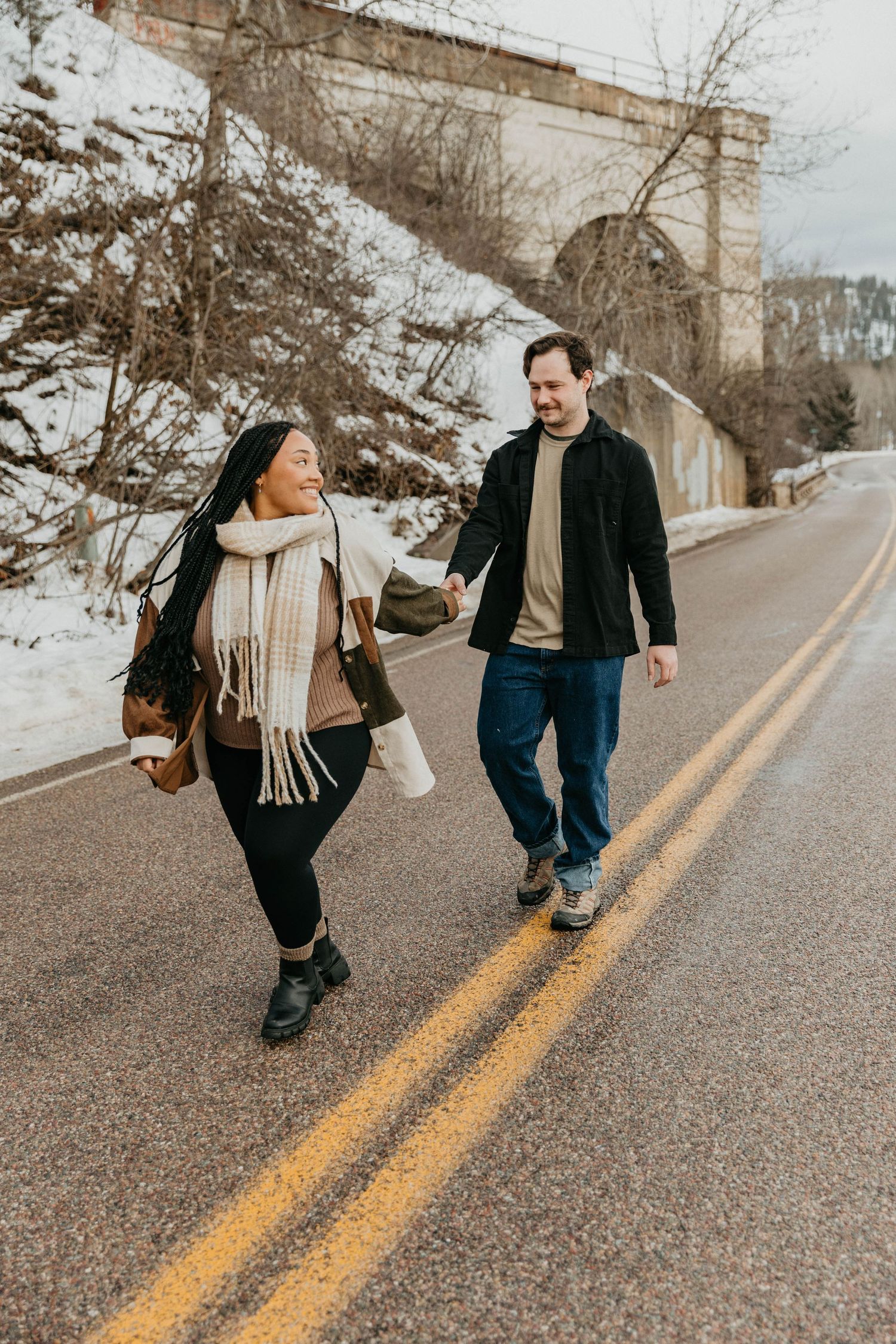 Two people walking together on a snowy road beneath a stone bridge.