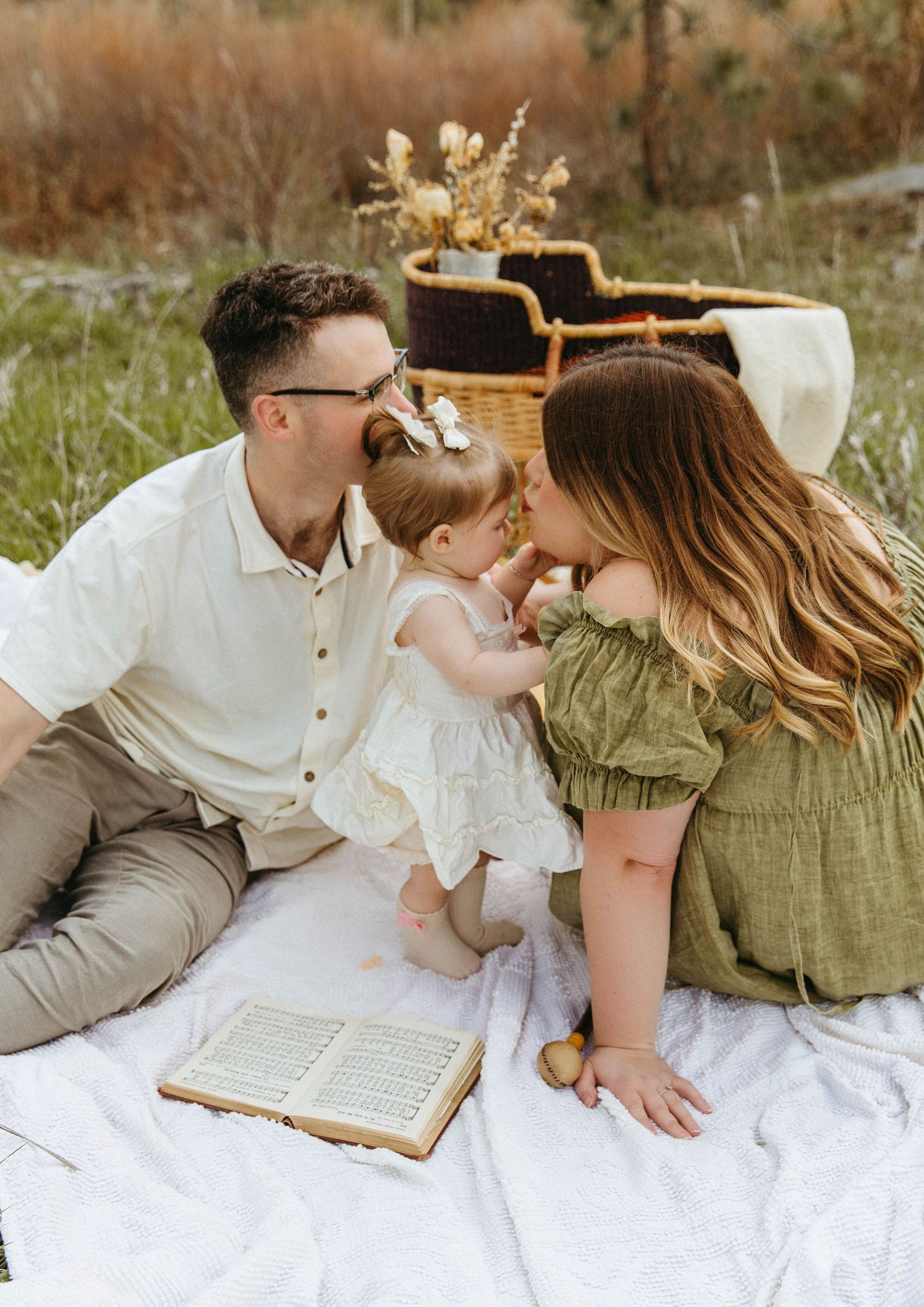 A family shares a tender moment together while sitting on a white blanket outdoors with dried flowers and an open book nearby.