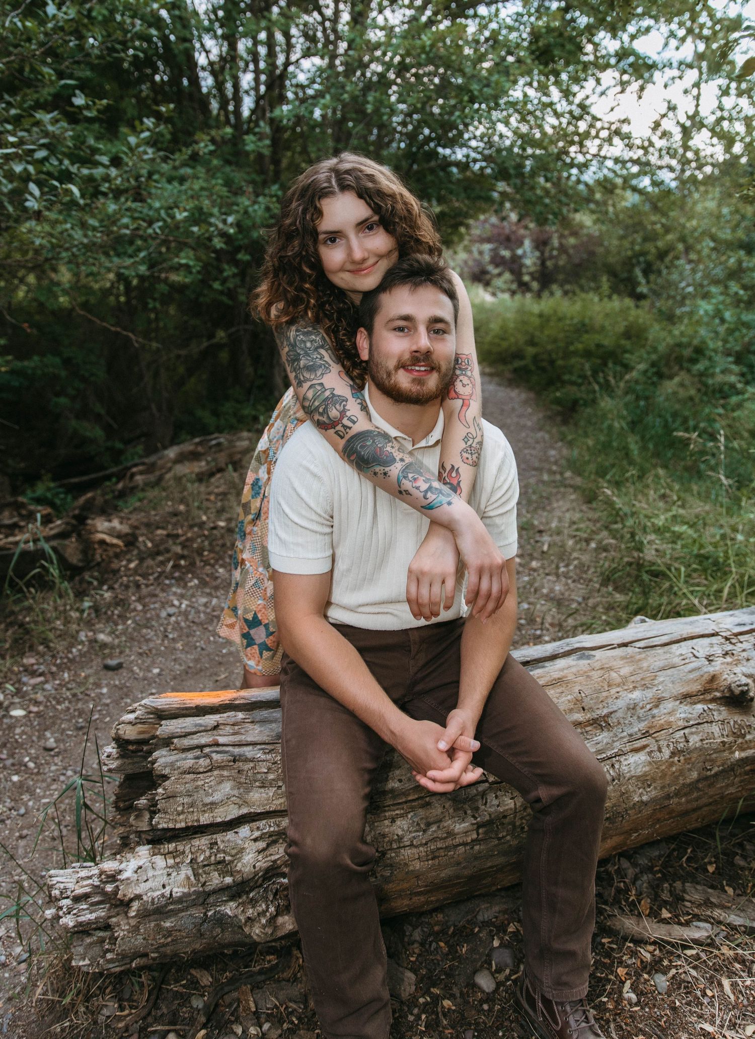 A couple poses for an intimate portrait while sitting on a fallen log in a wooded outdoor setting.