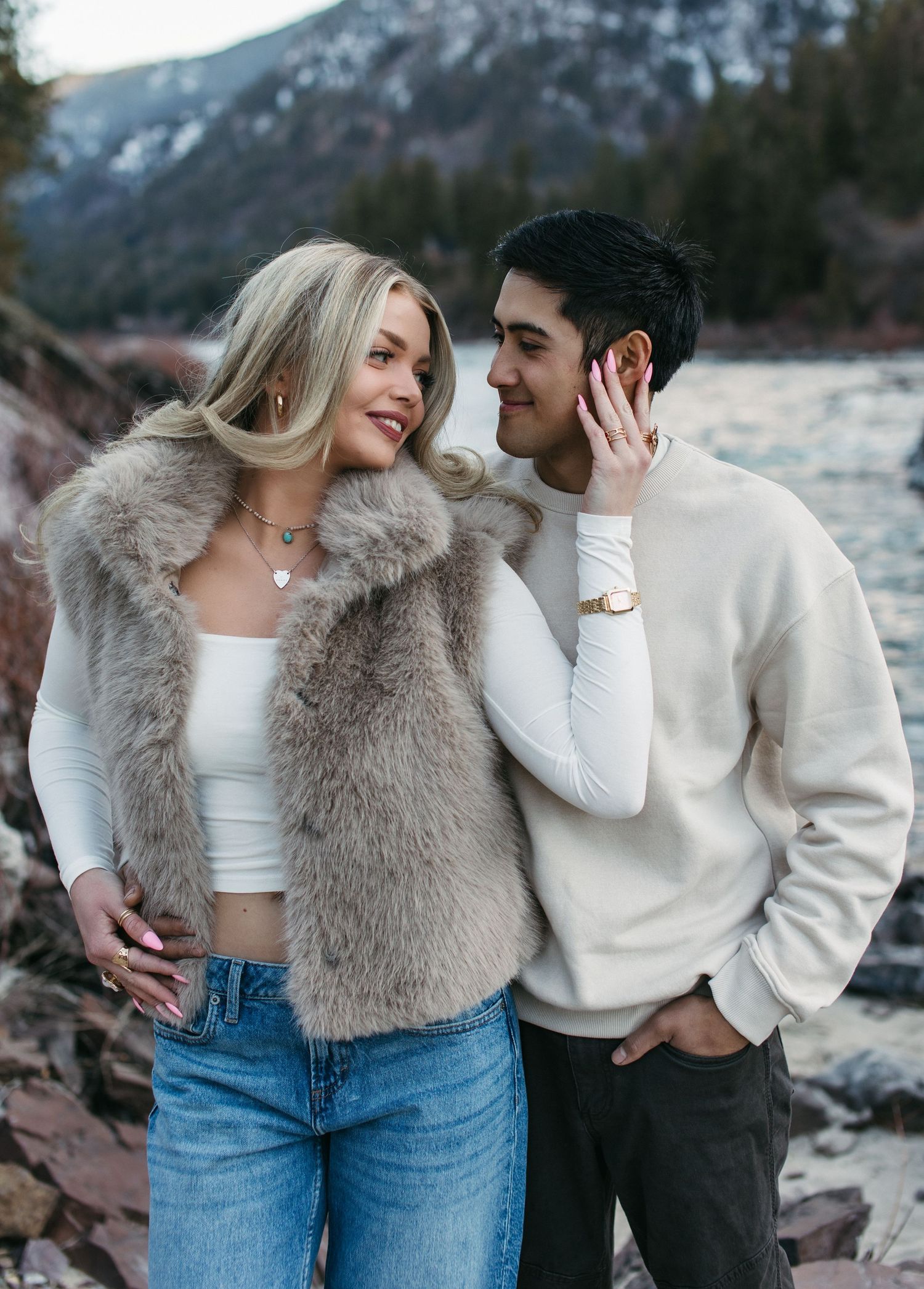 A couple in casual winter outfits embraces near a snowy mountain lake during a romantic photoshoot.