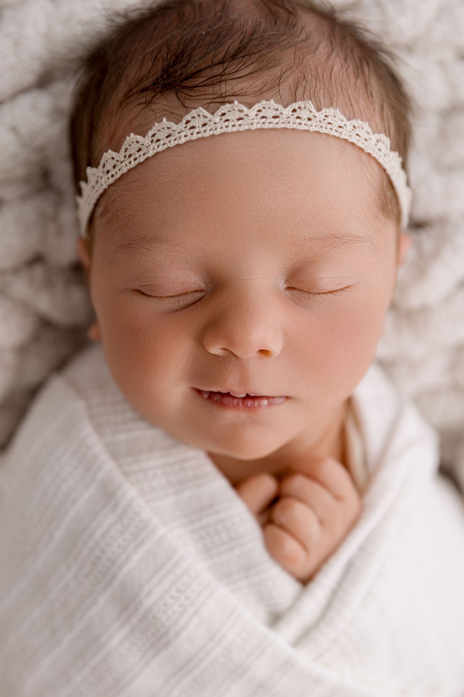 Sleeping newborn baby girl wrapped in white swaddle wearing a delicate lace headband, peaceful close-up portrait.