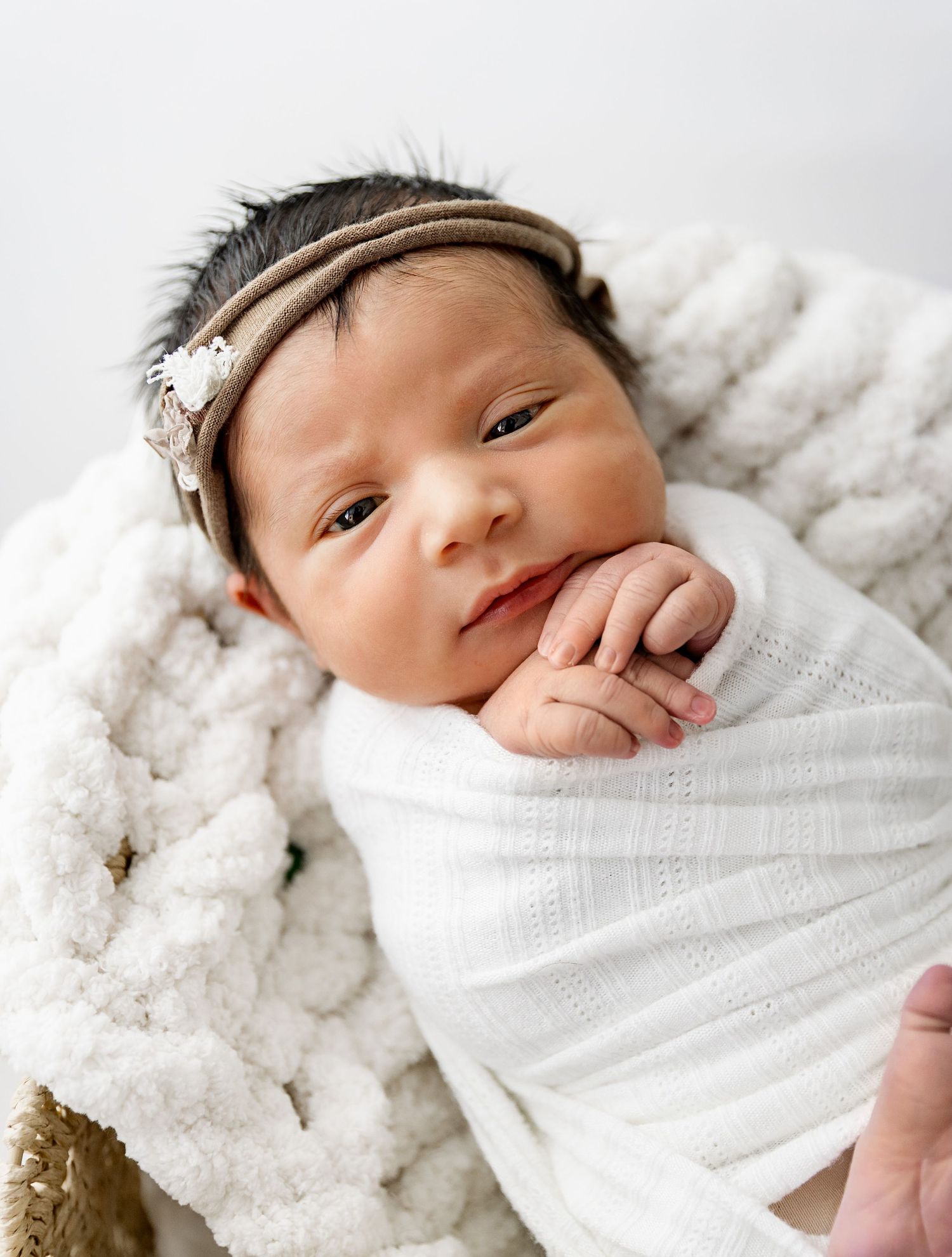 A newborn baby wrapped in white swaddle blanket rests peacefully on a textured white blanket.