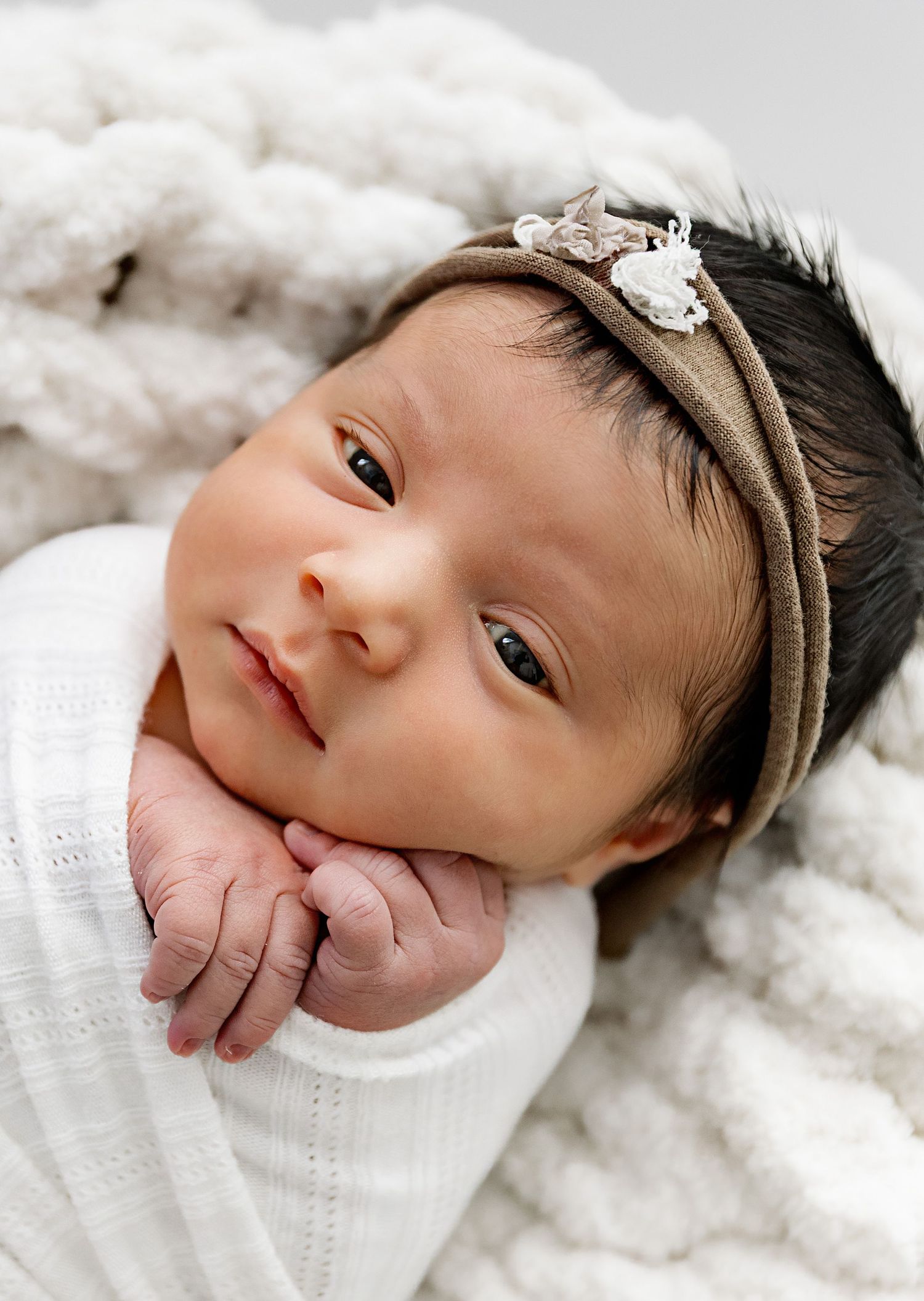 A baby wearing a white knit sweater and delicate floral headband rests on soft white blankets.