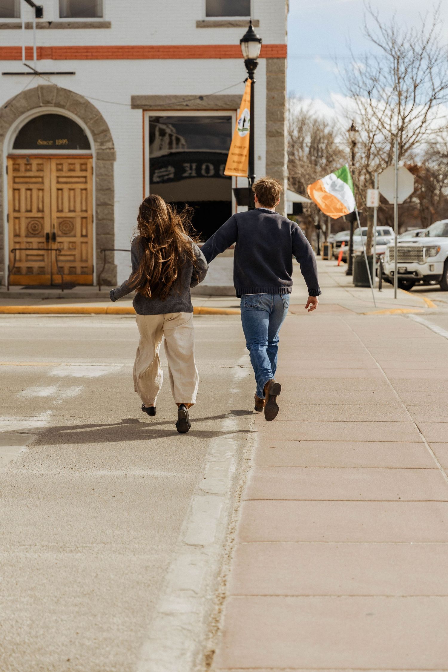 A couple walks together on a sunny sidewalk in front of a building with wooden doors.
