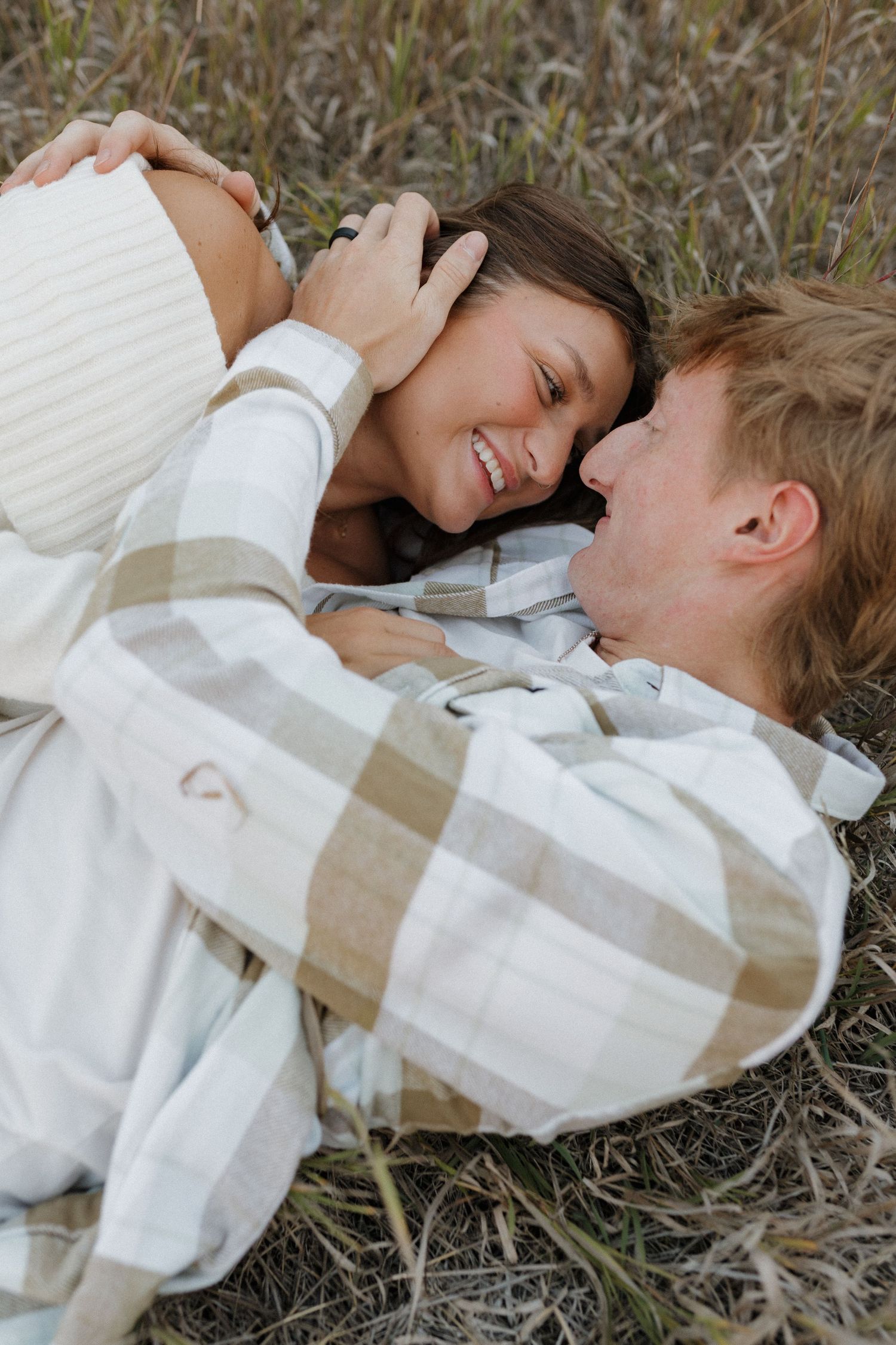 Two people in cozy white sweaters laughing while lying together on grass.