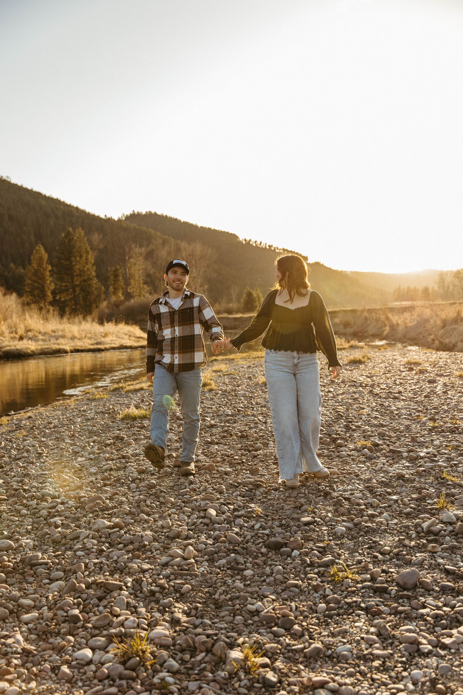 A couple walking hand in hand along a rocky riverbank at sunset.