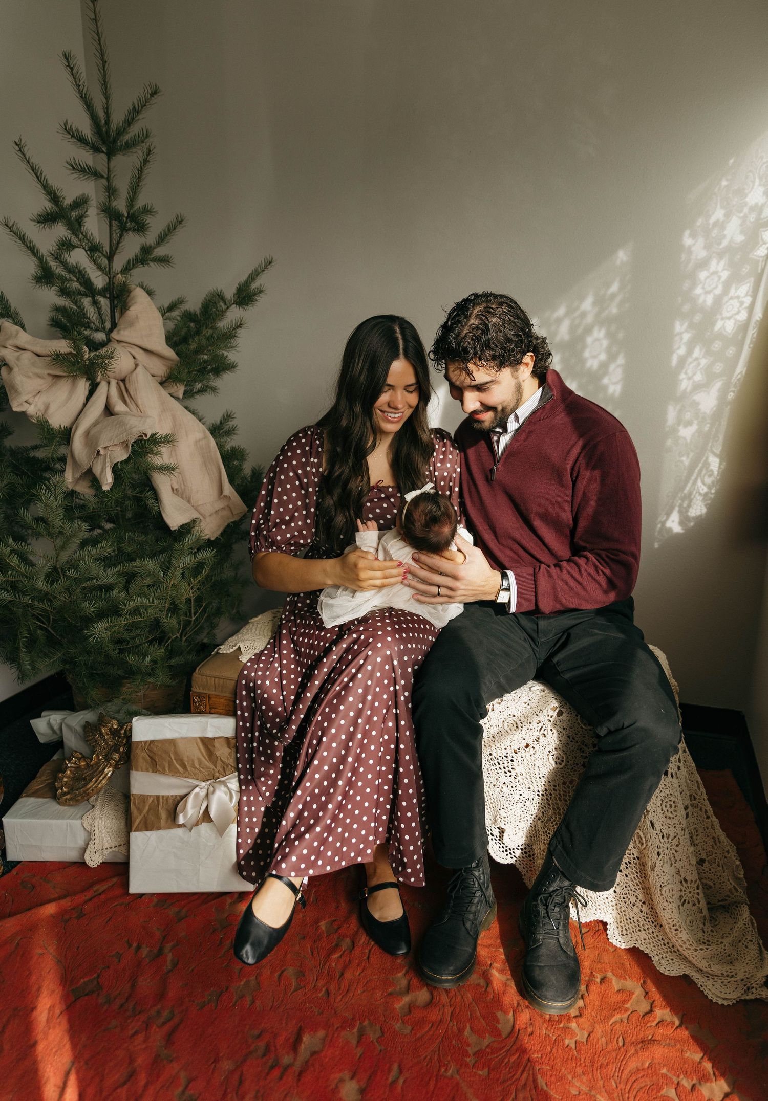 Couple in burgundy and polka dot dress share intimate moments on orange carpet near Christmas tree in vintage-style photoshoot.