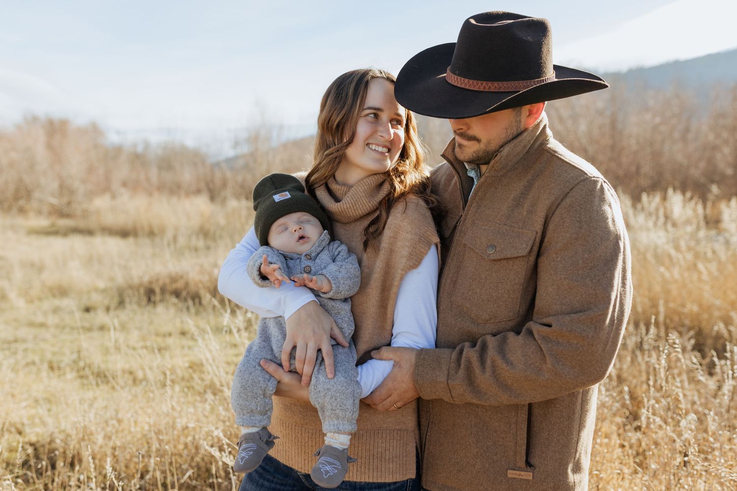 A couple holds their baby while sharing a tender moment in a sunlit field with mountains behind them.