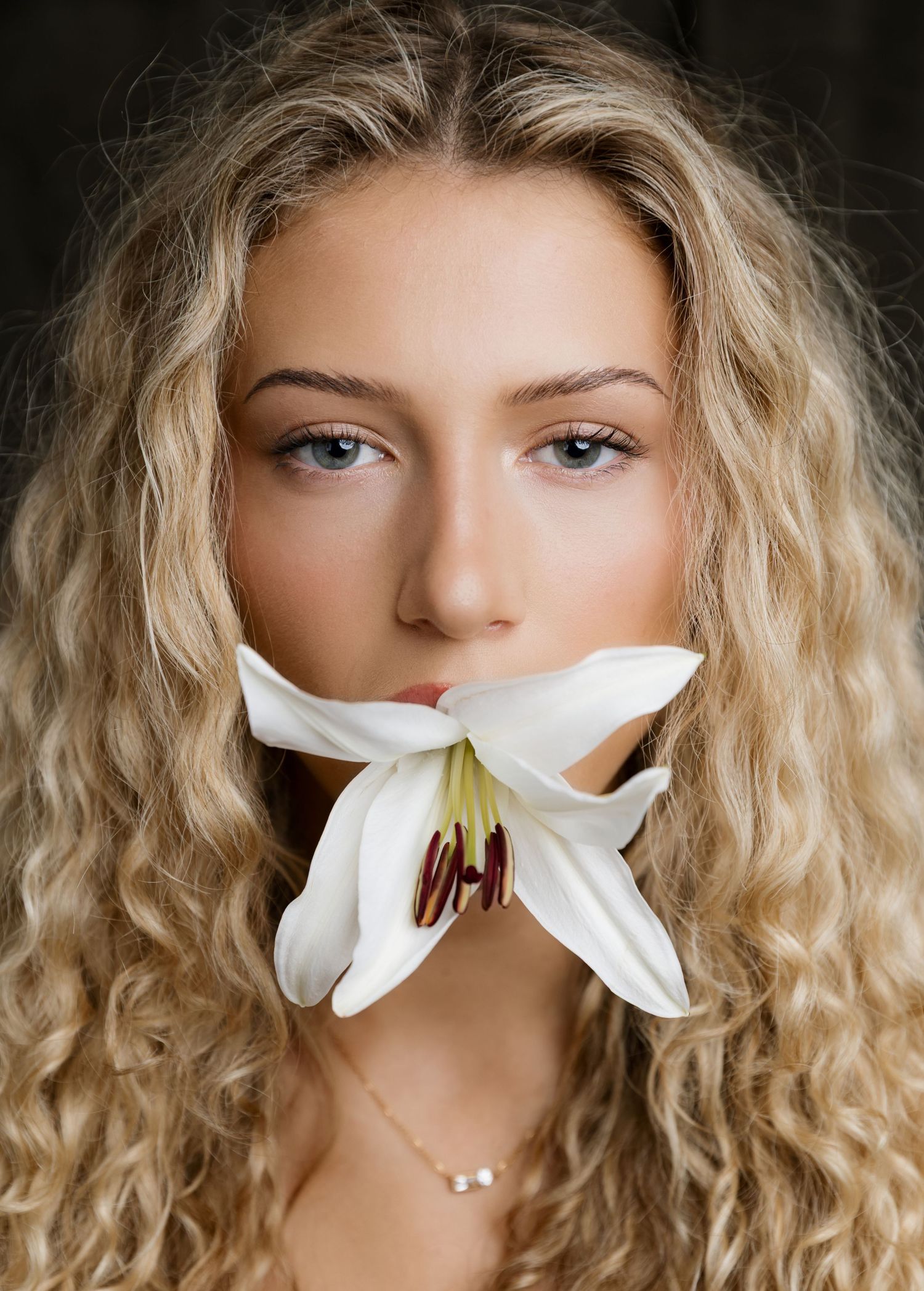 Young woman with curly blonde hair holding a white lily flower in her mouth against a dark background.