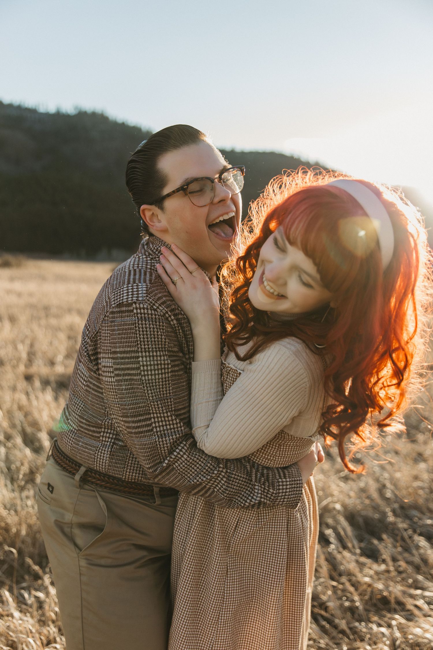 A joyful couple embraces in a golden field at sunset, the woman with red hair hugging a laughing man in vintage attire.