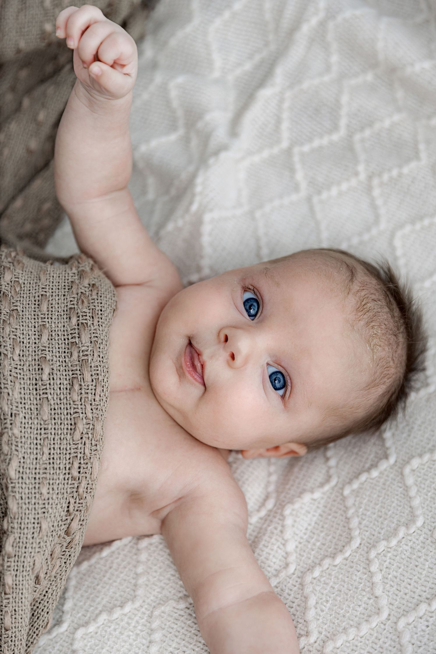 A baby with bright blue eyes lying on textured white bedding while looking up at the camera.