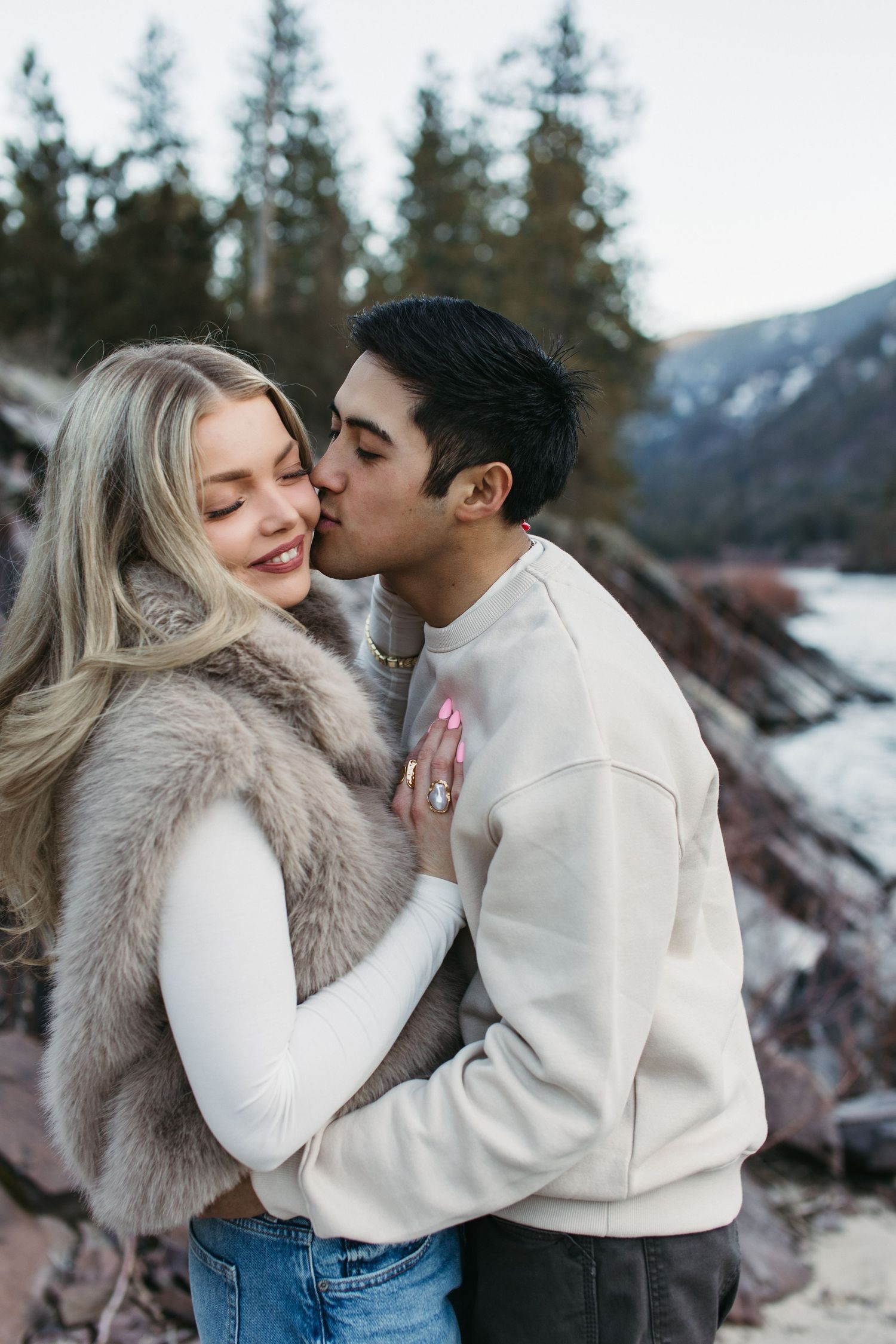 A romantic winter photoshoot by a snowy mountain lake with a couple in matching cream sweaters sharing intimate moments.