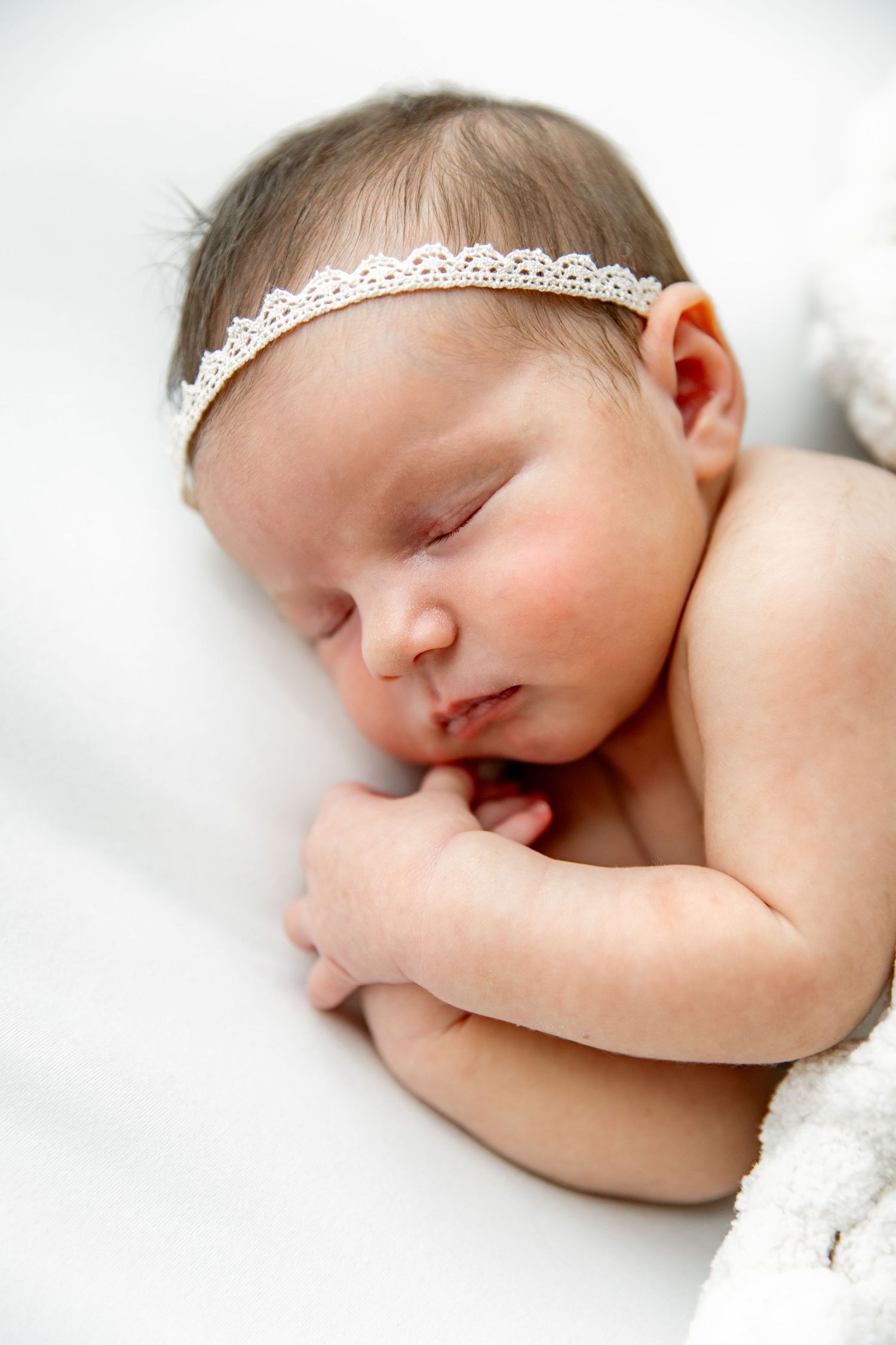 A sleeping newborn baby wearing a delicate lace headband rests peacefully on soft white bedding.