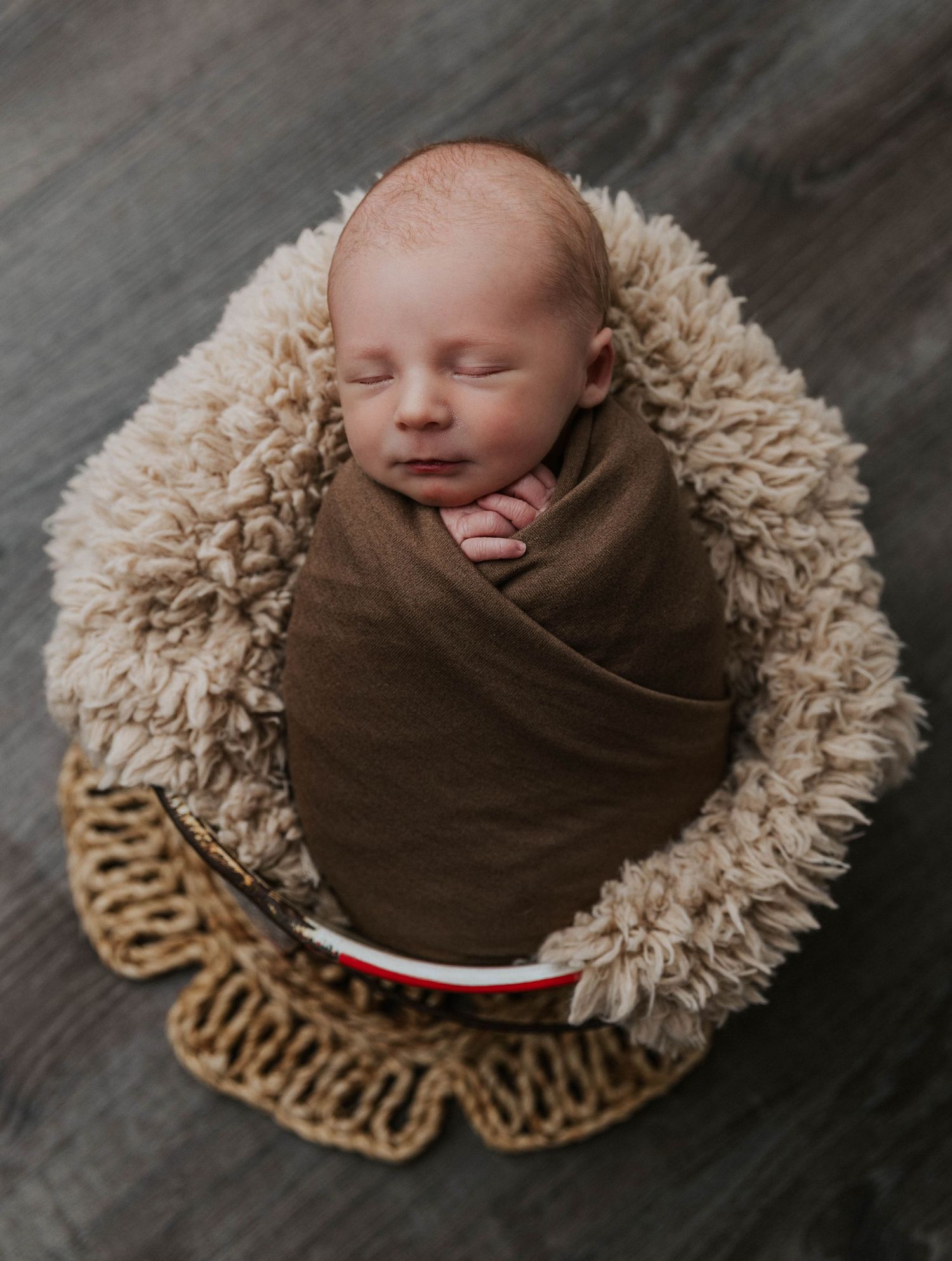 A newborn wrapped in a brown swaddle rests on a textured cream-colored fur blanket with woven decor underneath.