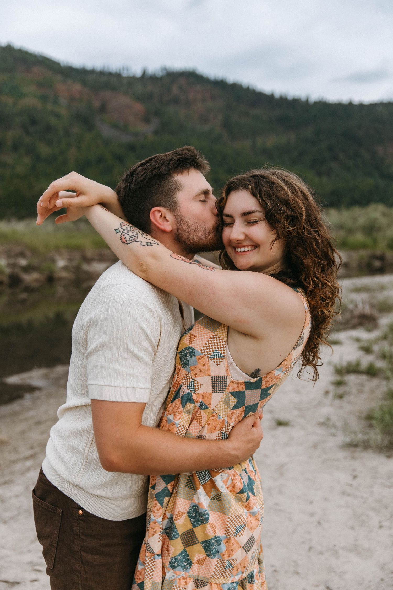 A couple shares tender moments while embracing on a rocky beach with mountains in the background.
