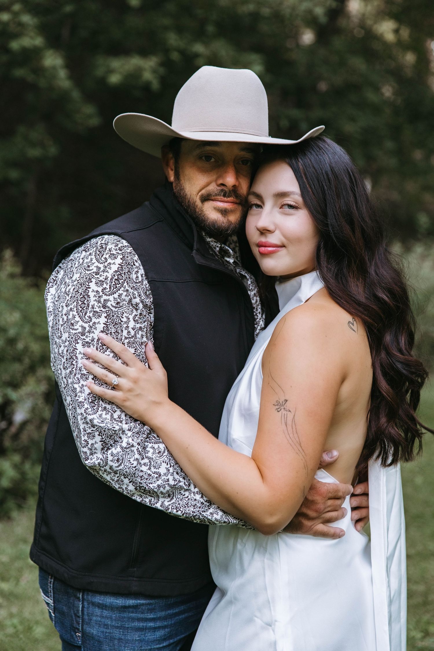A romantic couple in western attire embraces outdoors during a photo session in a natural setting.