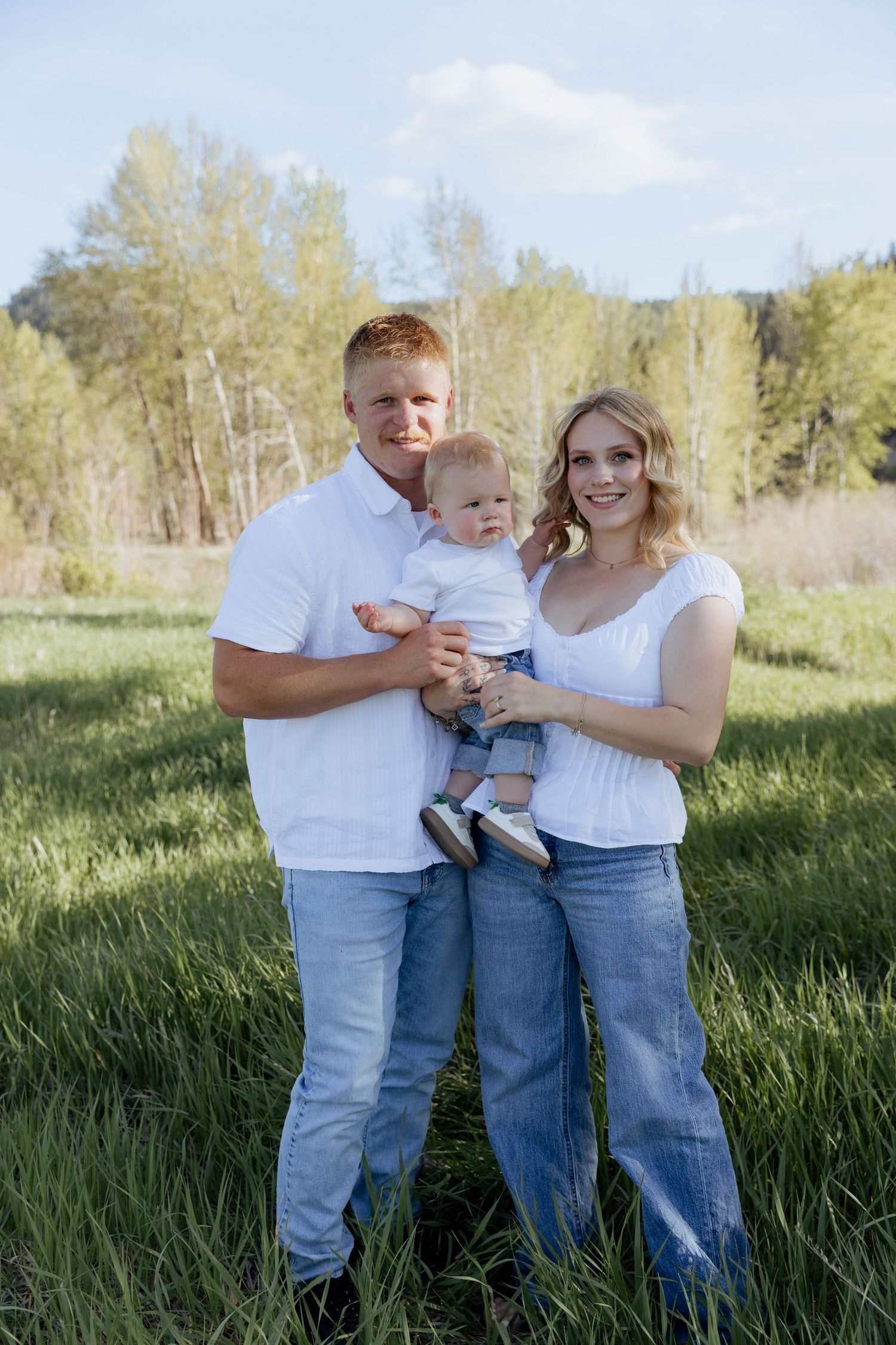 Young family in white tops and jeans posing outdoors in green meadow with trees, parents holding baby.
