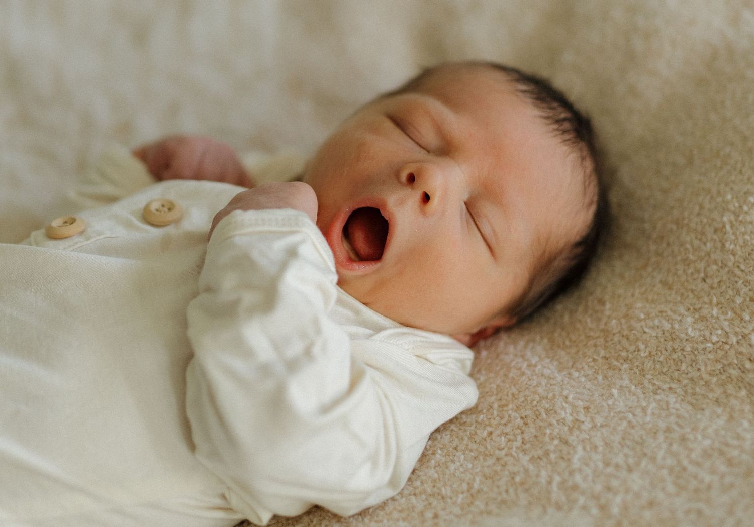 Sleeping baby in white onesie yawning while lying on cream colored surface.