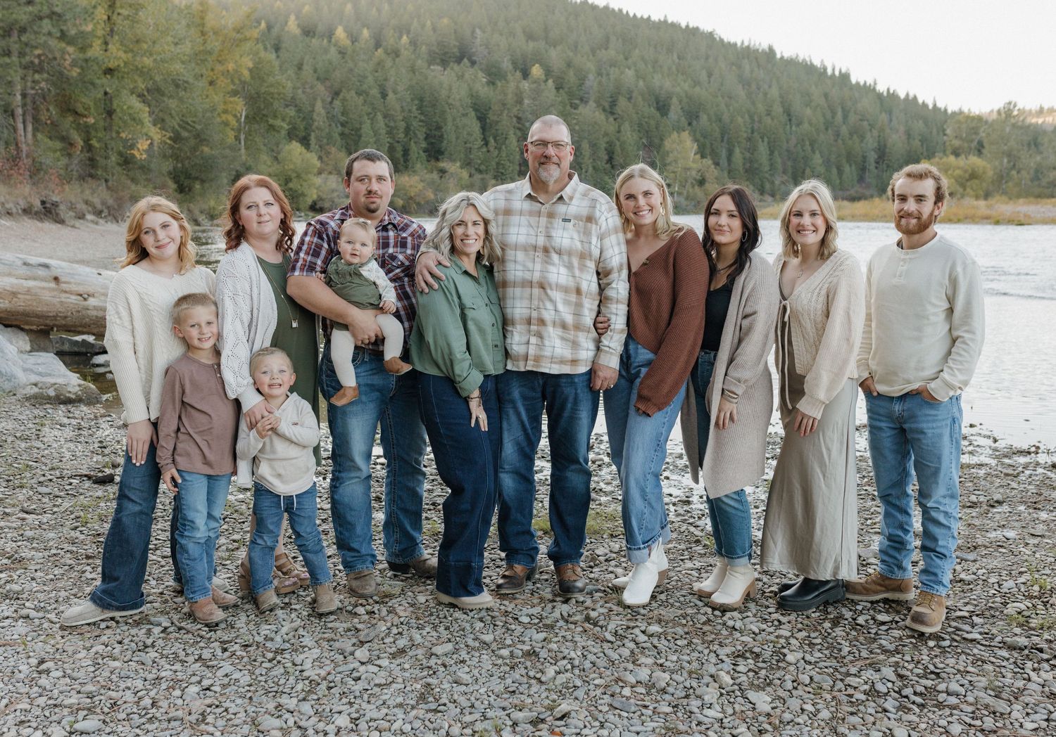 Extended family group photo on rocky beach with evergreen forest and mountains in background.