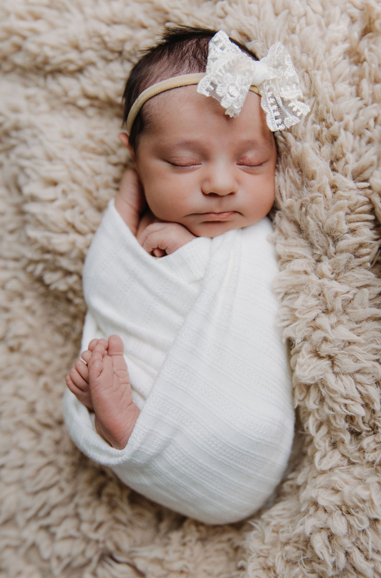 Sleeping newborn in white swaddle with lace bow headband on fluffy beige blanket.