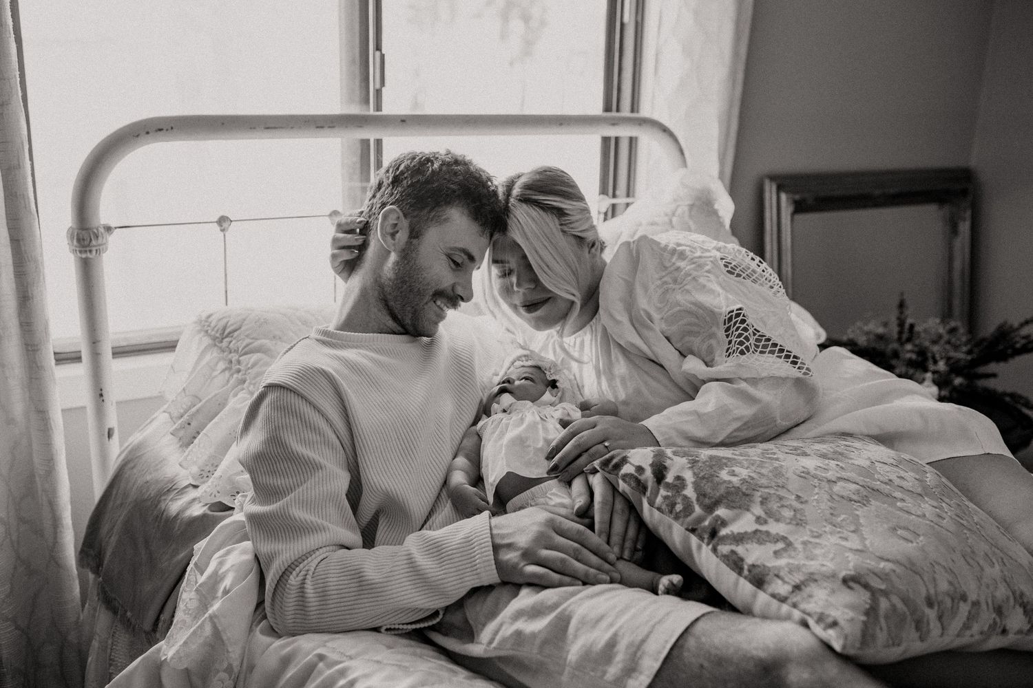 An intimate black and white photograph of a couple with their newborn on a bed.