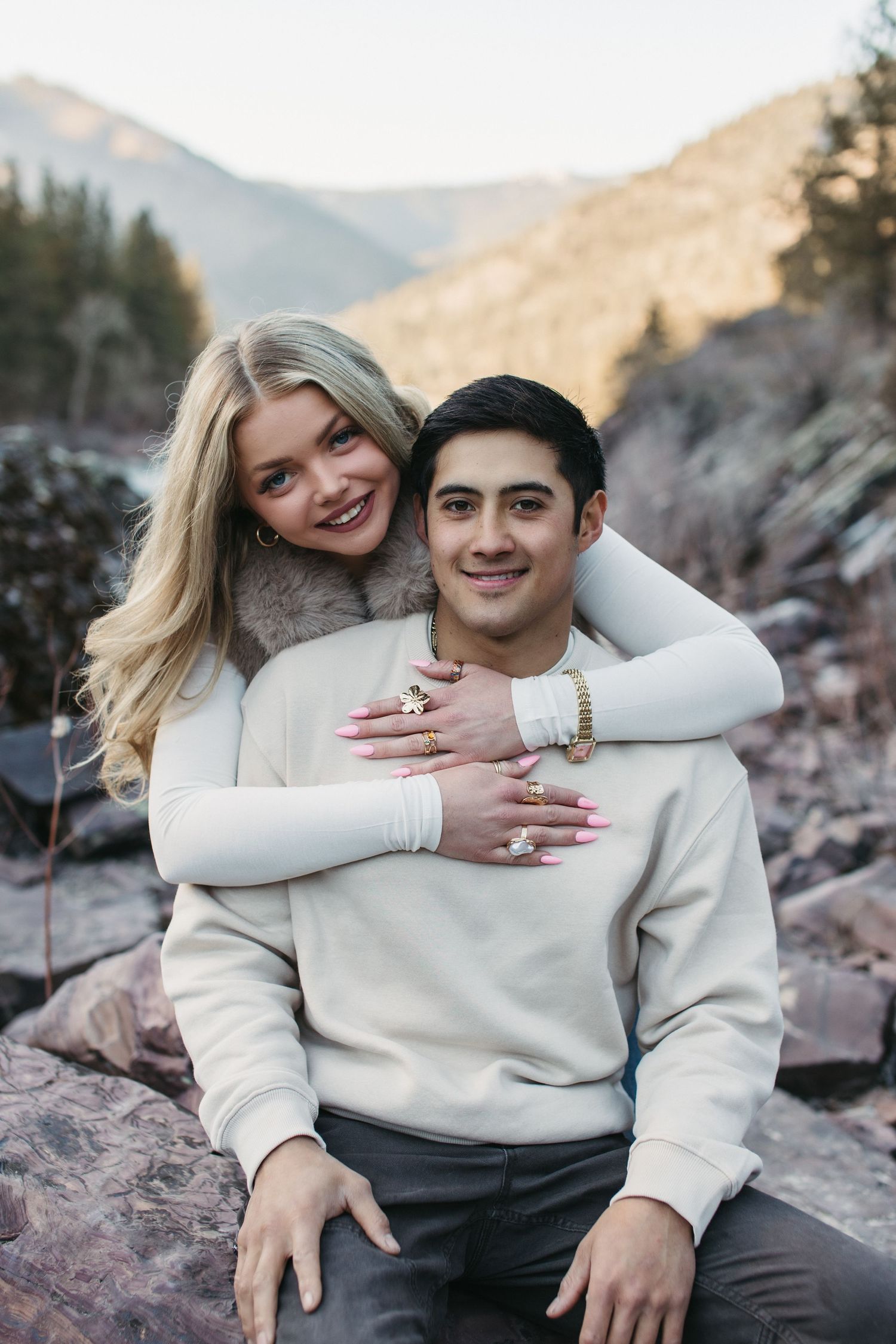 A couple in matching cream sweaters embrace on rocky mountain terrain during a romantic engagement photo session.