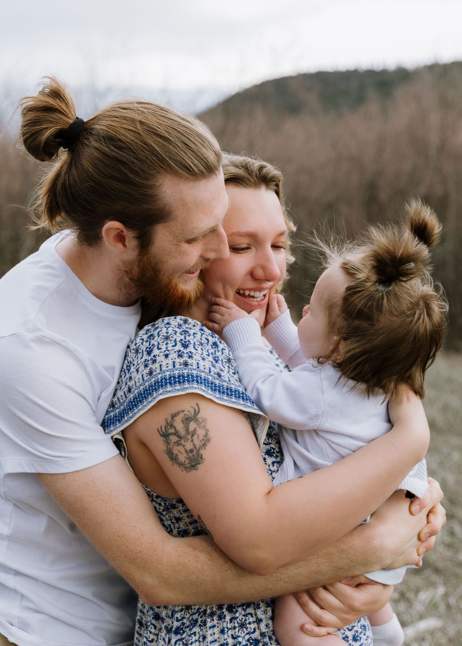 A happy young family with tattoos embraces outdoors, parents kissing and hugging their toddler daughter in nature.