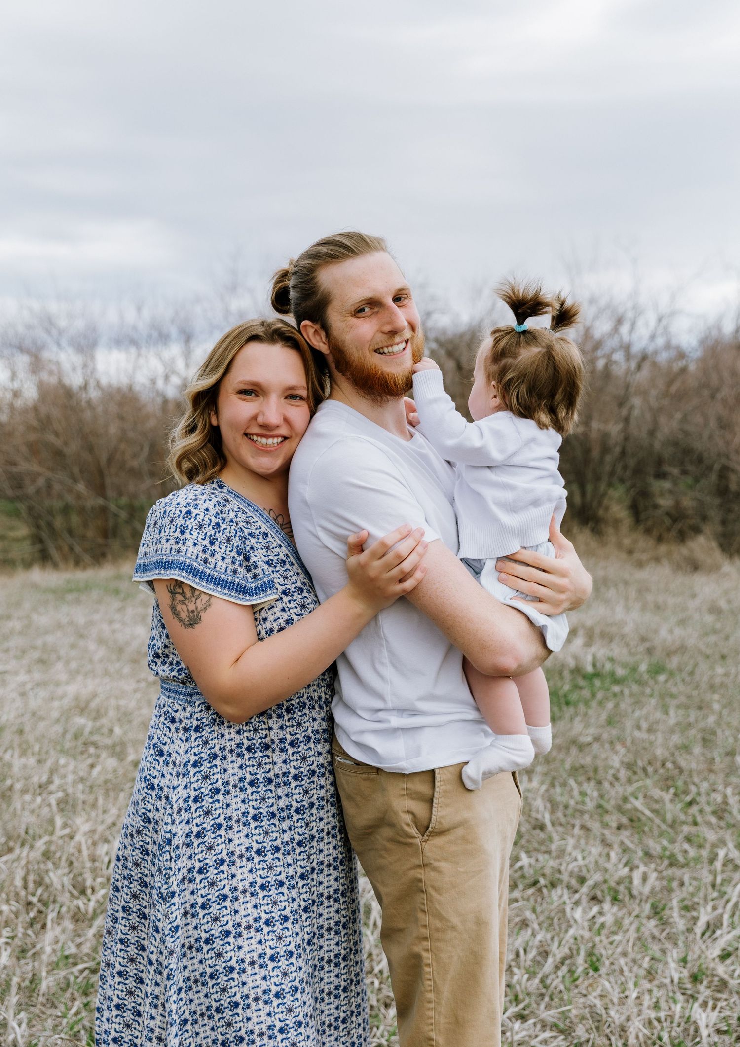 Young family smiling outdoors in a field, man holding toddler girl while woman in blue dress embraces them.