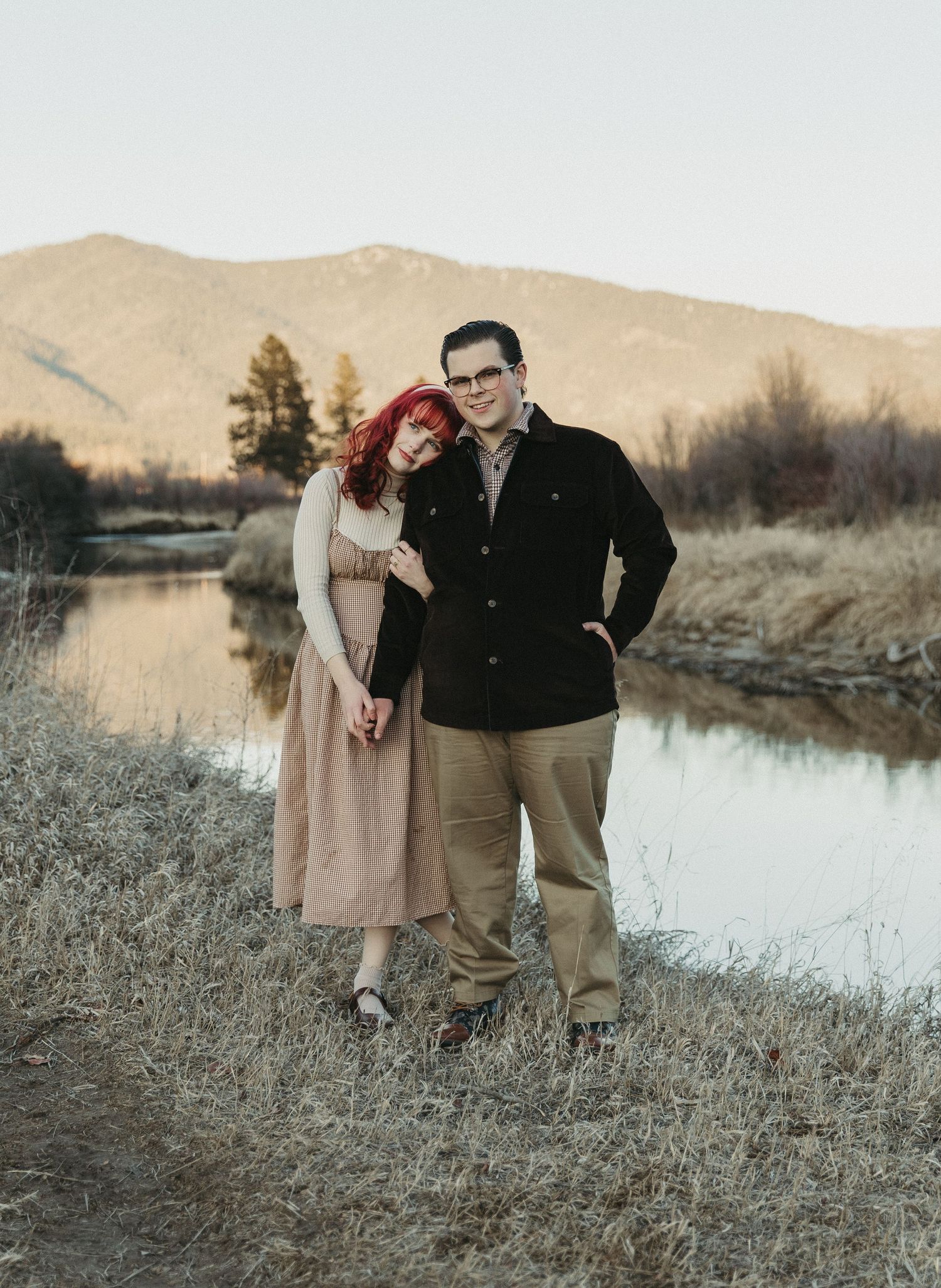 A couple poses by a mountain river, woman in pink dress with red hair leans on man in black jacket amid dry grass.