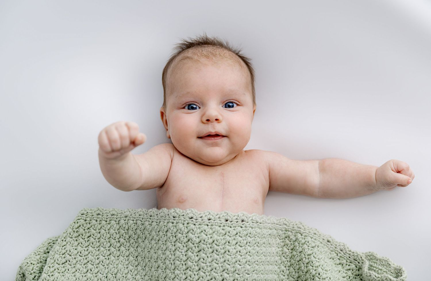 Baby lying on white bedding covered partially with mint green knitted blanket.