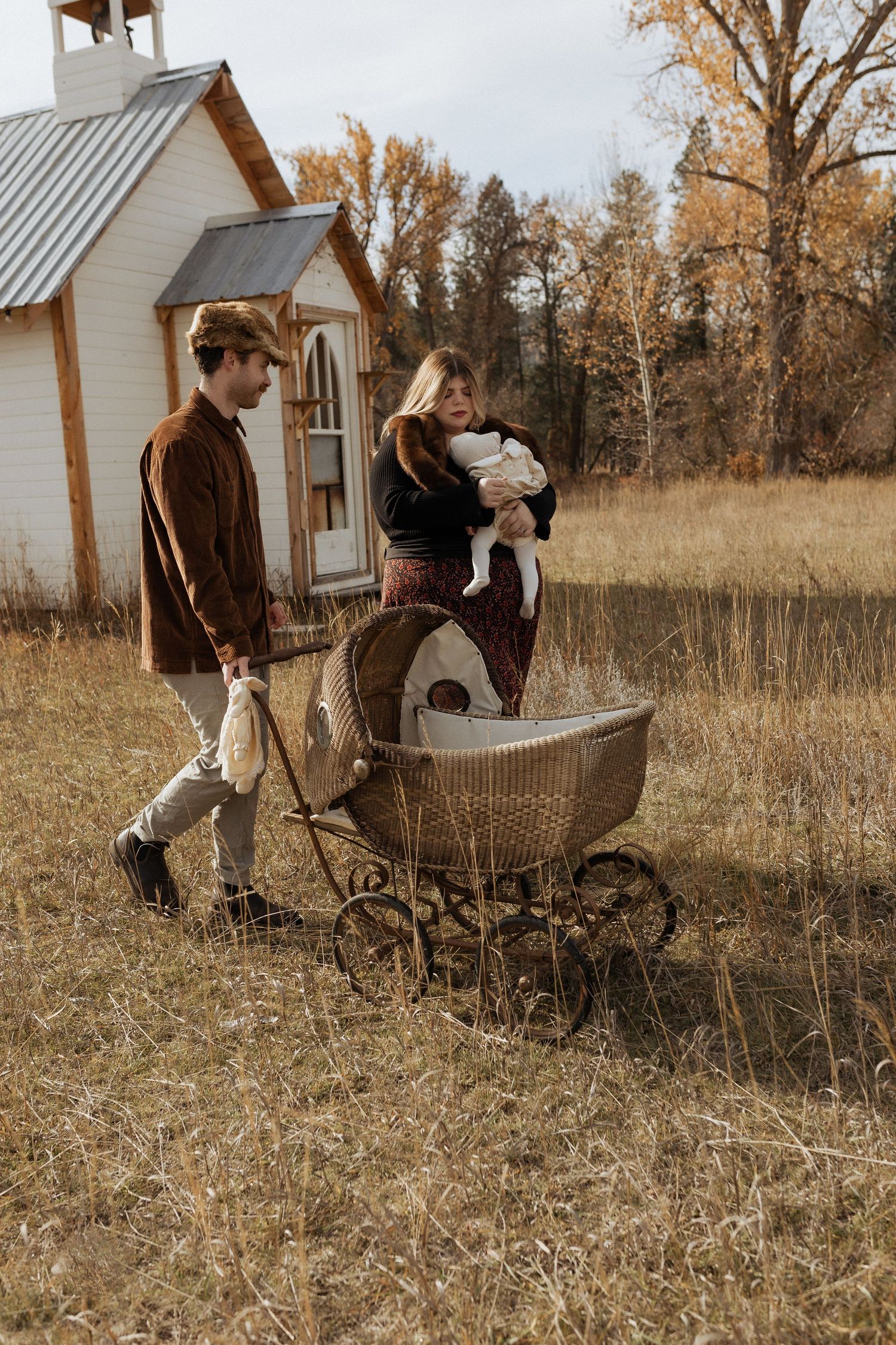 Family walking with vintage pram near rustic white chapel in autumn field.