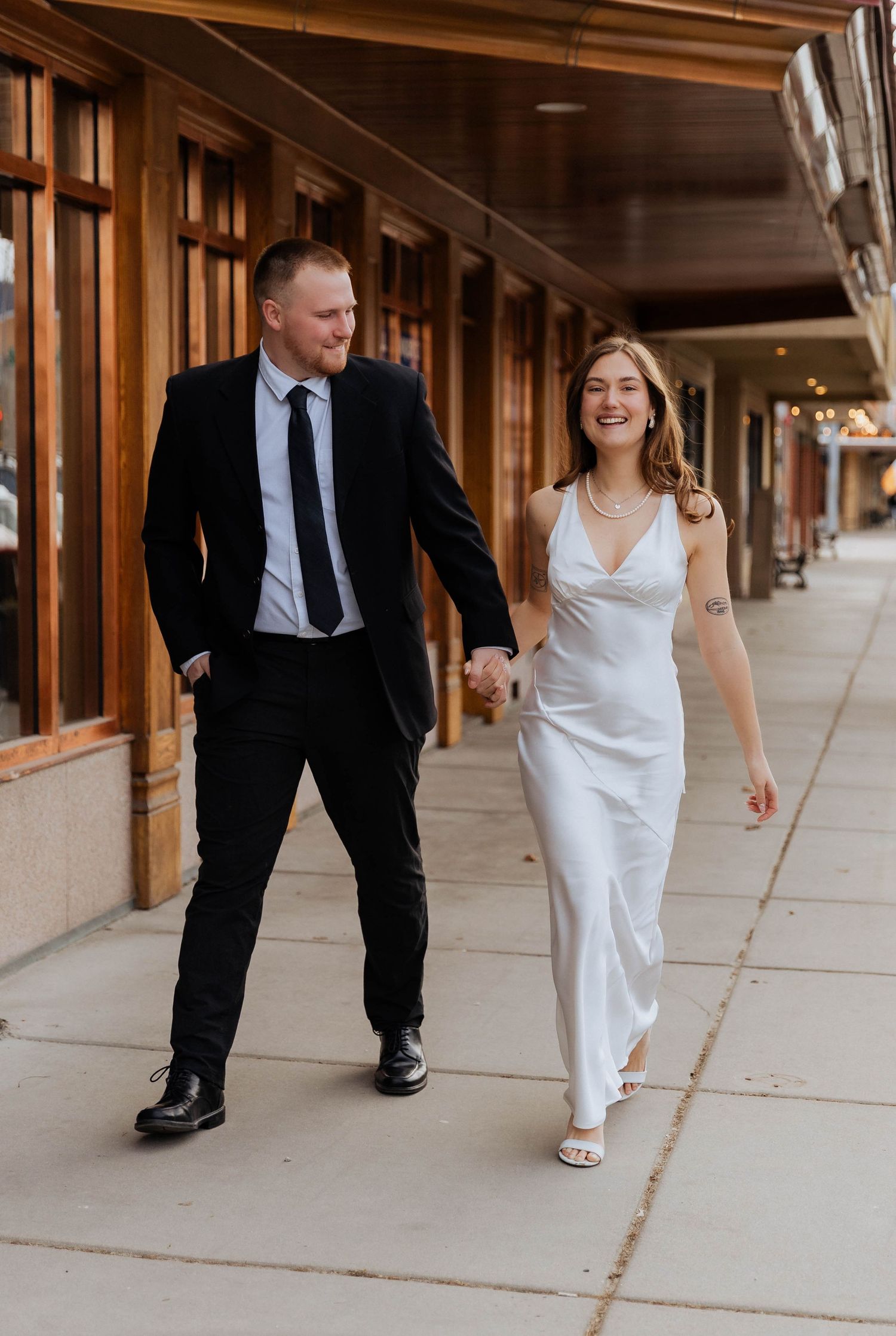 Couple in formal attire walking and laughing together on a downtown sidewalk.