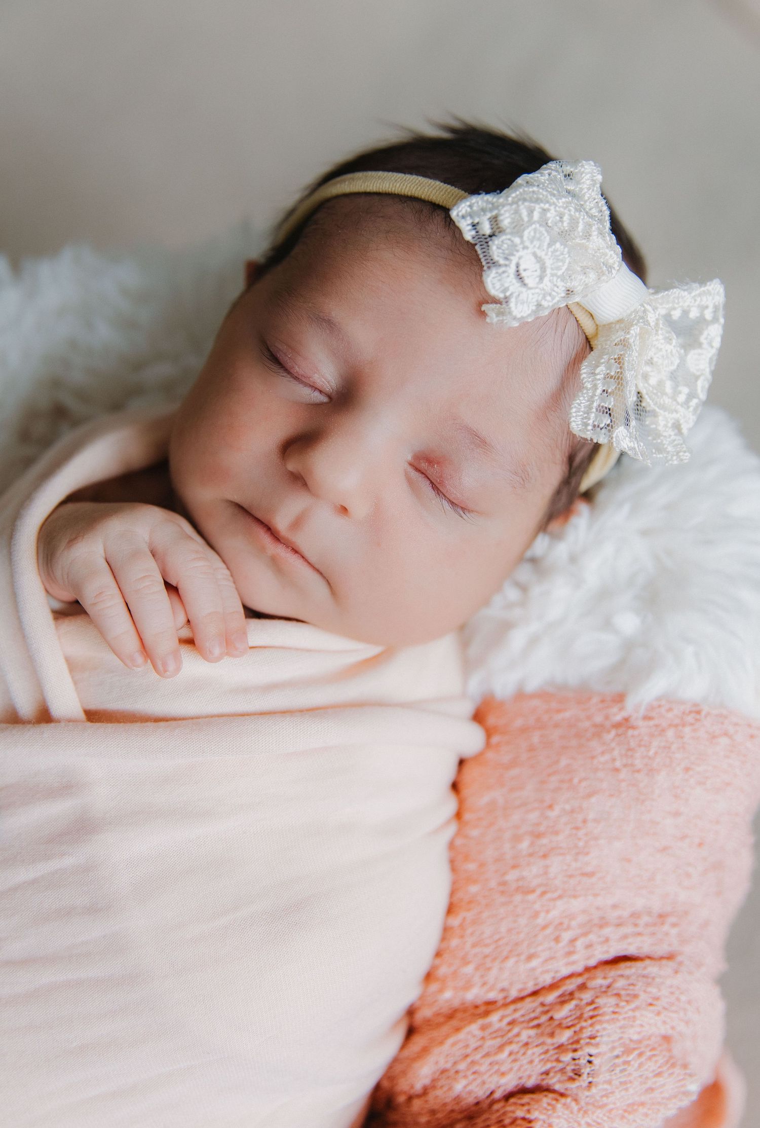 Newborn sleeping peacefully in pink swaddle with white lace headband on soft blankets.