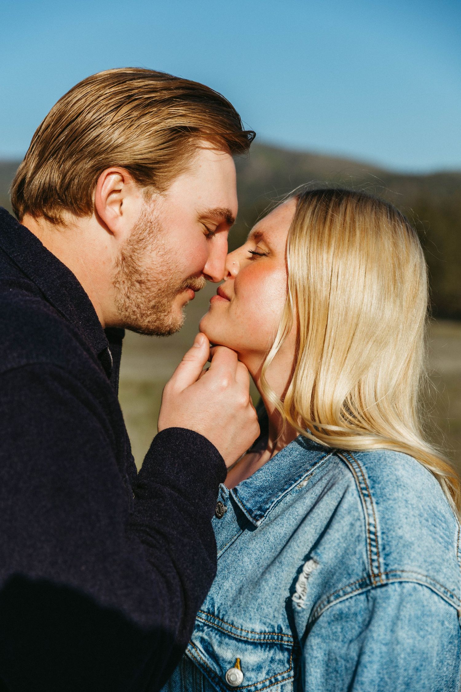 A romantic couple sharing an intimate kiss outdoors while wearing denim and dark clothing on a sunny day.