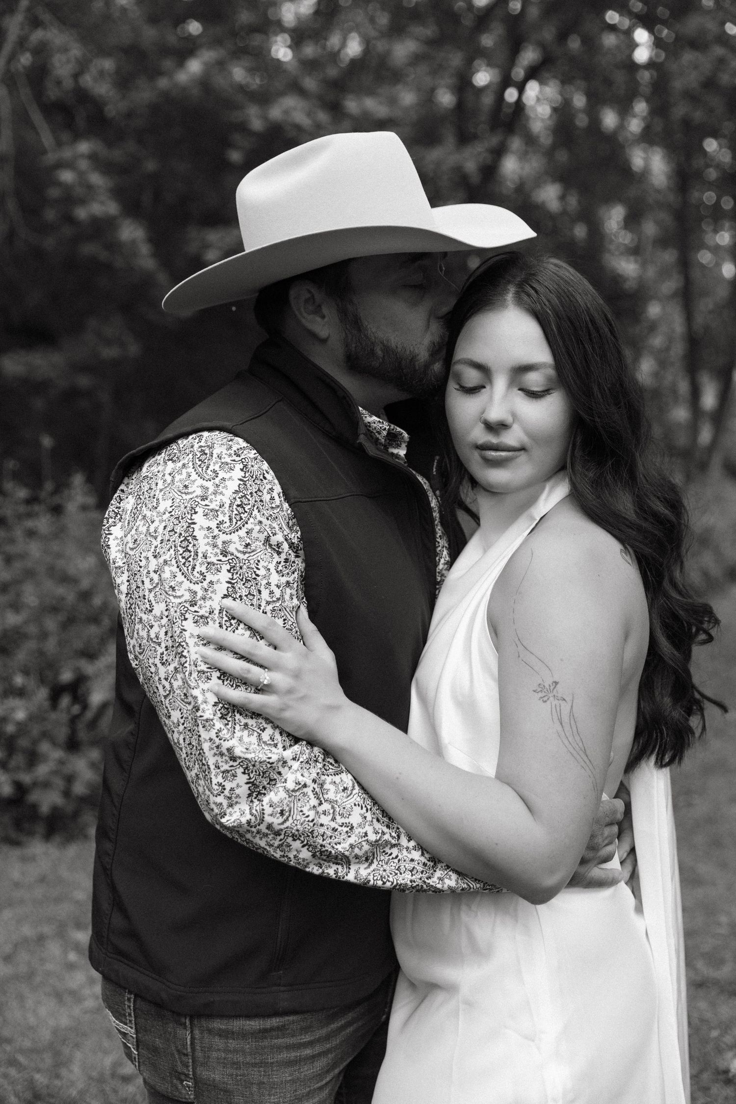 A romantic black and white photo of a couple embracing outdoors, with one wearing a cowboy hat and sequined shirt.