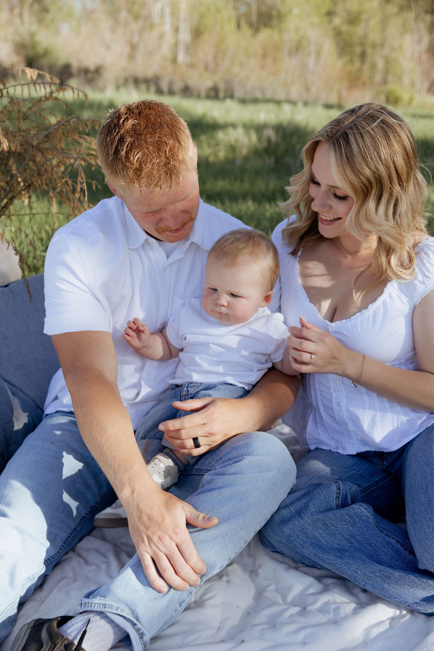 A happy young family with a red-haired father, blonde mother, and baby in white outfits sitting on a blanket outdoors.