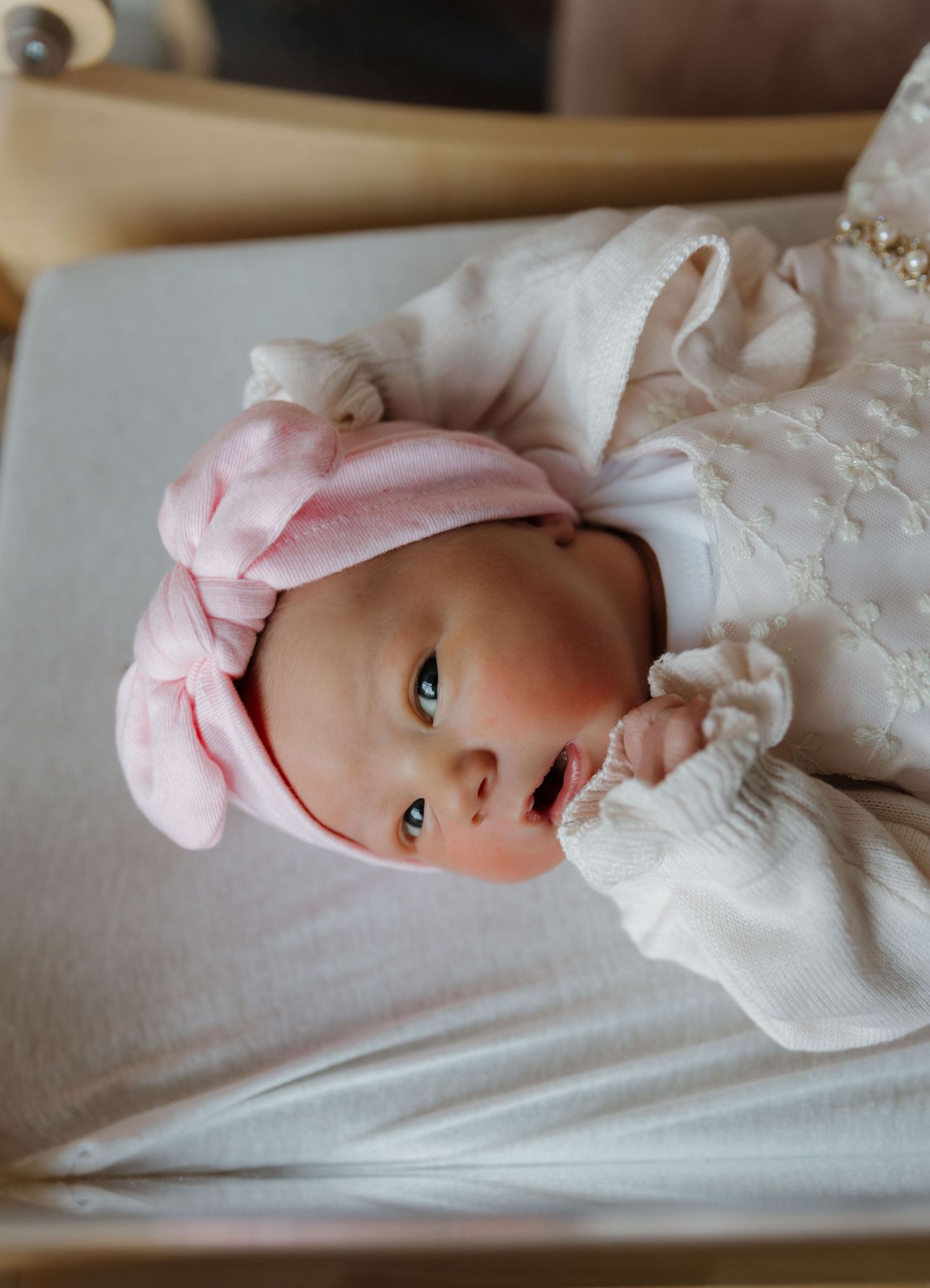 Newborn in white dress and pink bow headband resting on light colored bedding.